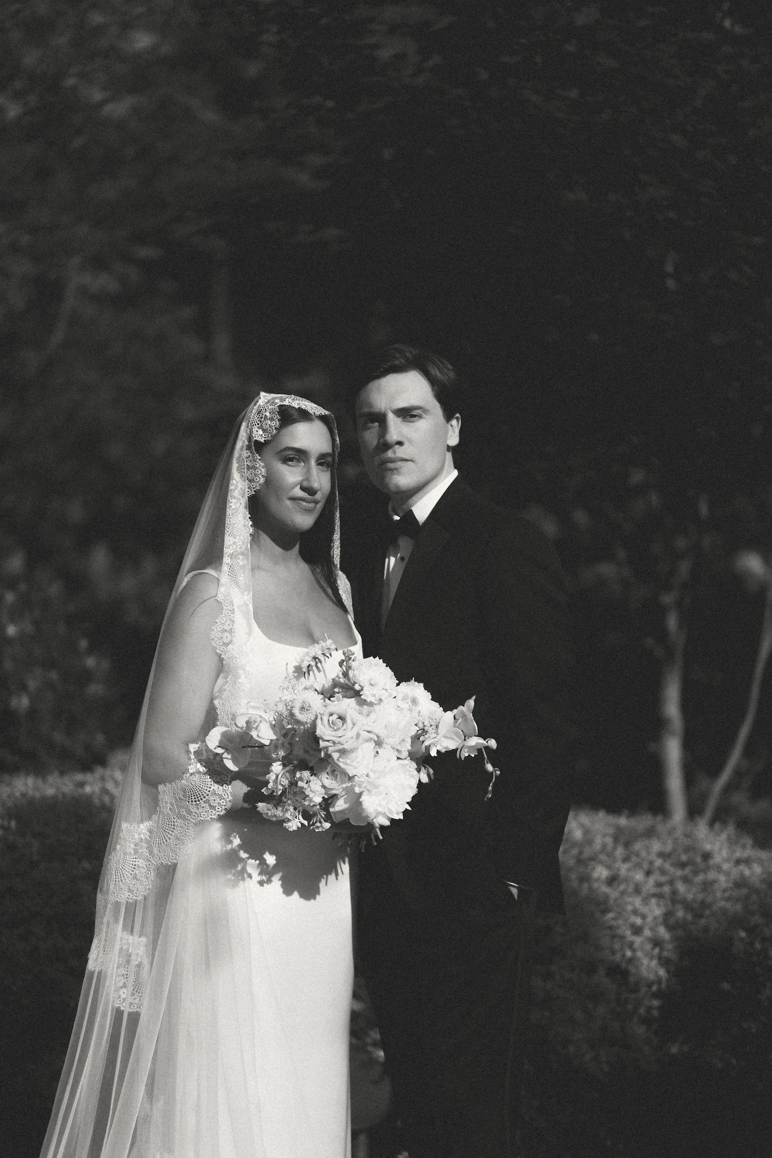 Black and white photo of a bride and groom standing outdoors at night with trees in the background. The bride is wearing a wedding dress and veil and holding a bouquet of flowers. The groom is dressed in a tuxedo.