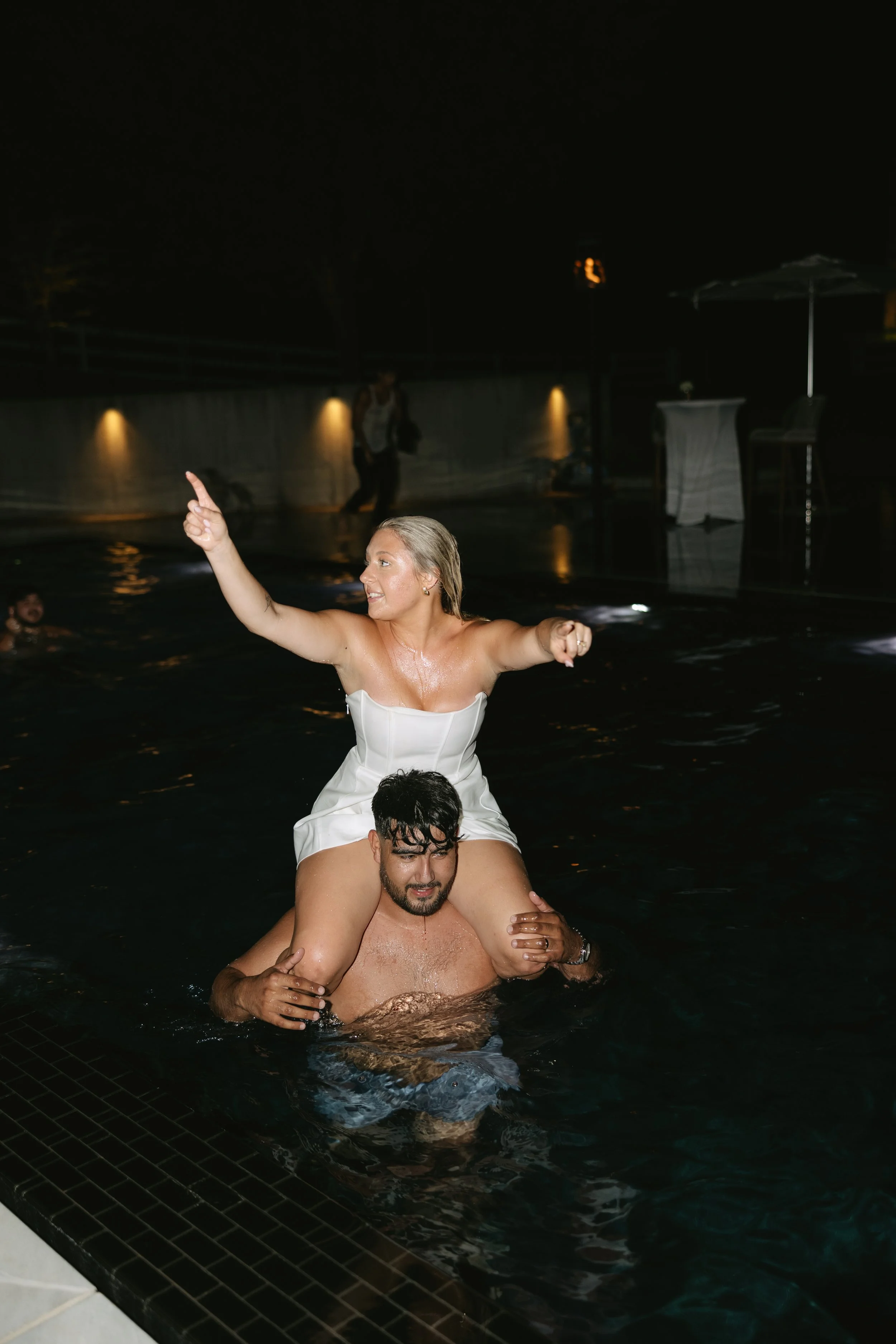 A woman in a white dress sitting on a man's shoulders in a pool during a nighttime event.