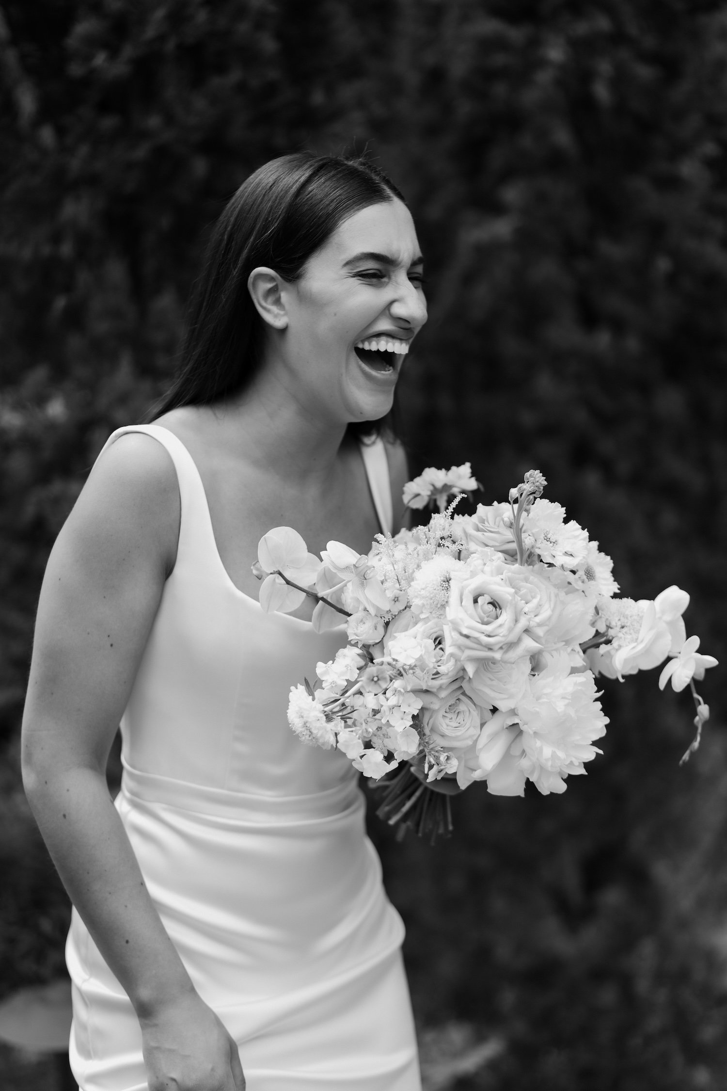 A woman in a white dress holding a large bouquet of flowers, laughing outdoors with a blurred background.