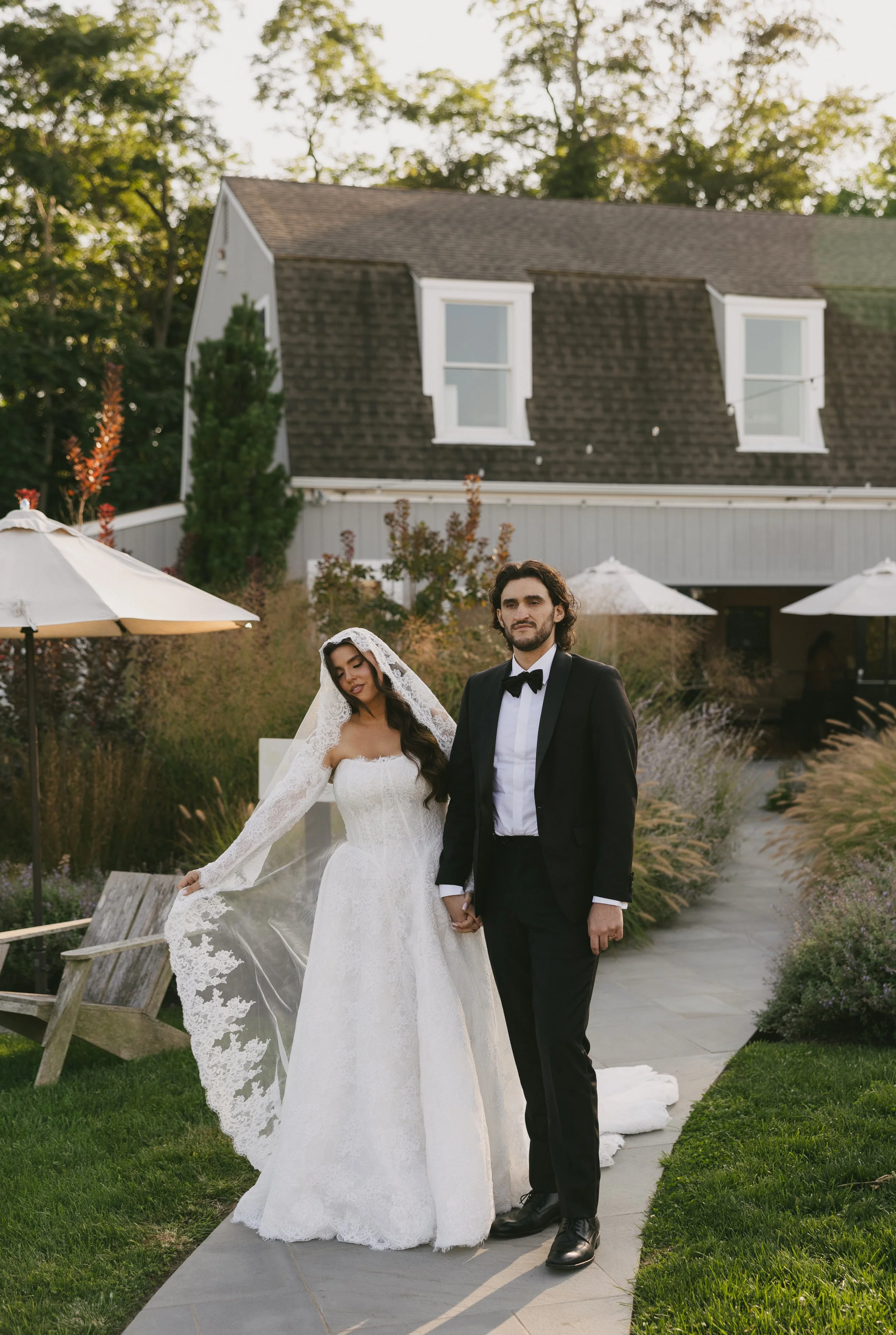 A bride and groom holding hands outdoors, with the bride in a white wedding gown and lace veil, and the groom in a black tuxedo with a bowtie, standing on a concrete path in a garden.