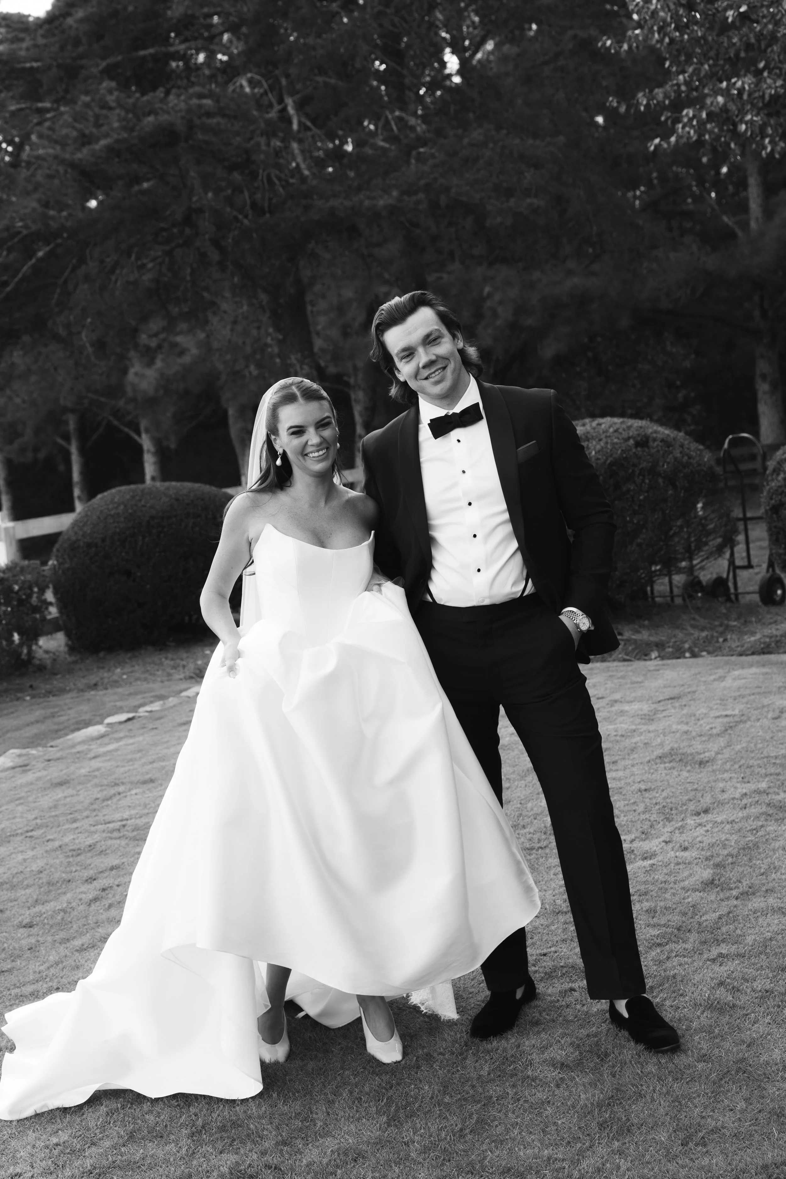Black and white photo of a bride and groom standing together outdoors, smiling, with trees and bushes in the background. The bride is wearing a strapless wedding gown and the groom is in a tuxedo with a bow tie.