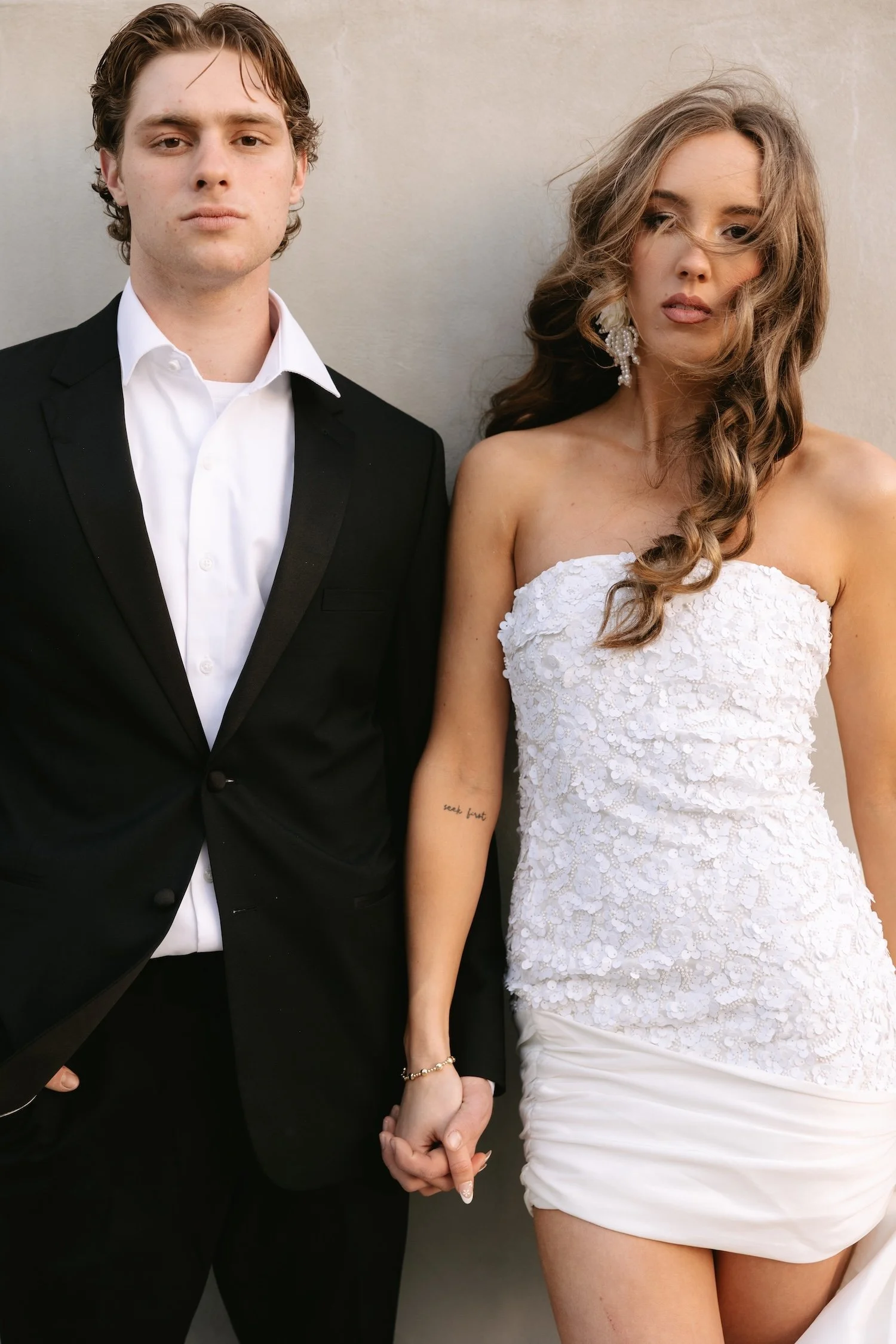 A young man and woman, dressed in formal attire, standing side-by-side and holding hands. The man is wearing a black tuxedo with a white shirt, and the woman is wearing a strapless white dress with lace details. The woman has wavy hair and is wearing