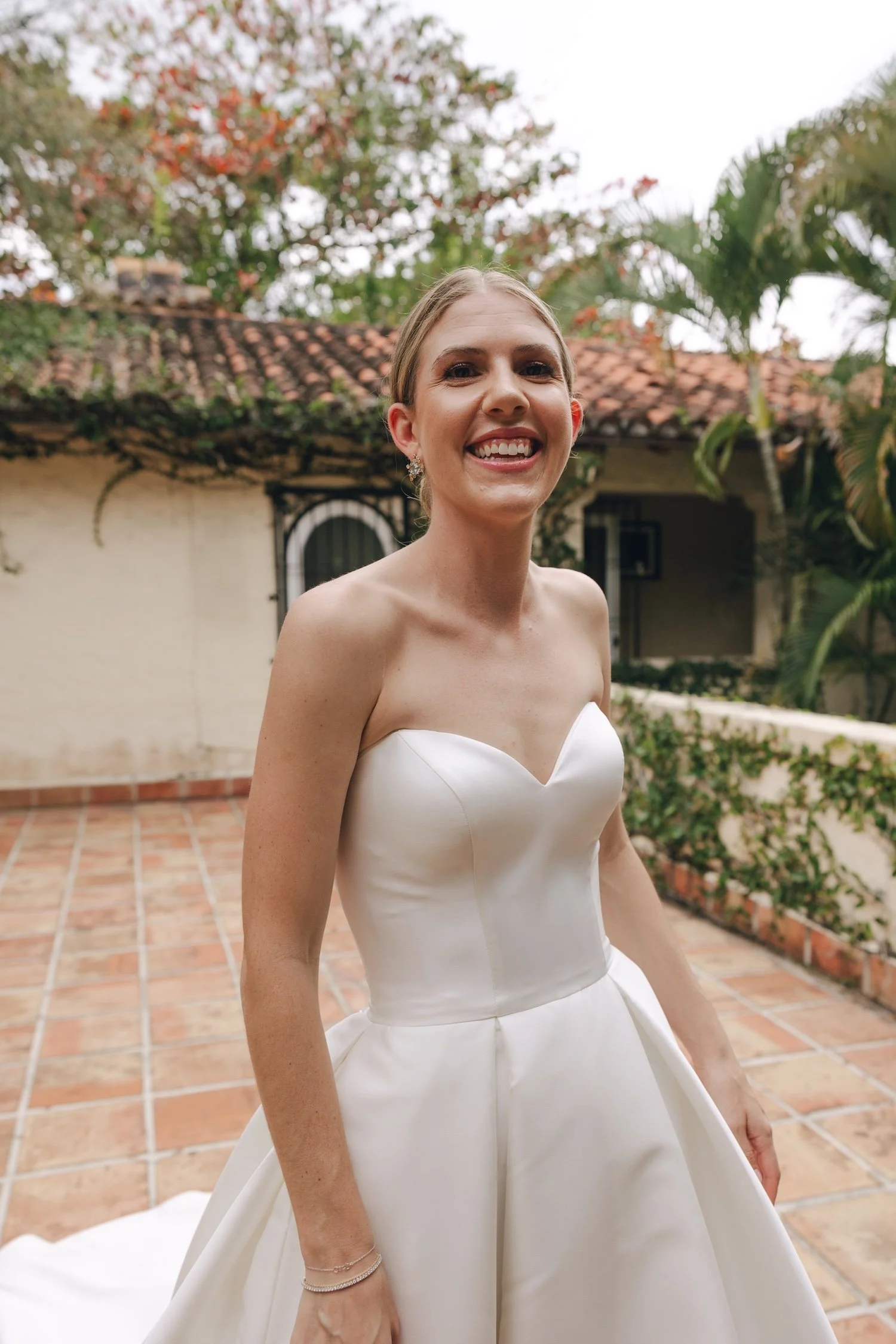 A smiling bride in a strapless white wedding gown standing outdoors on a tiled patio, with a garden and old building in the background.