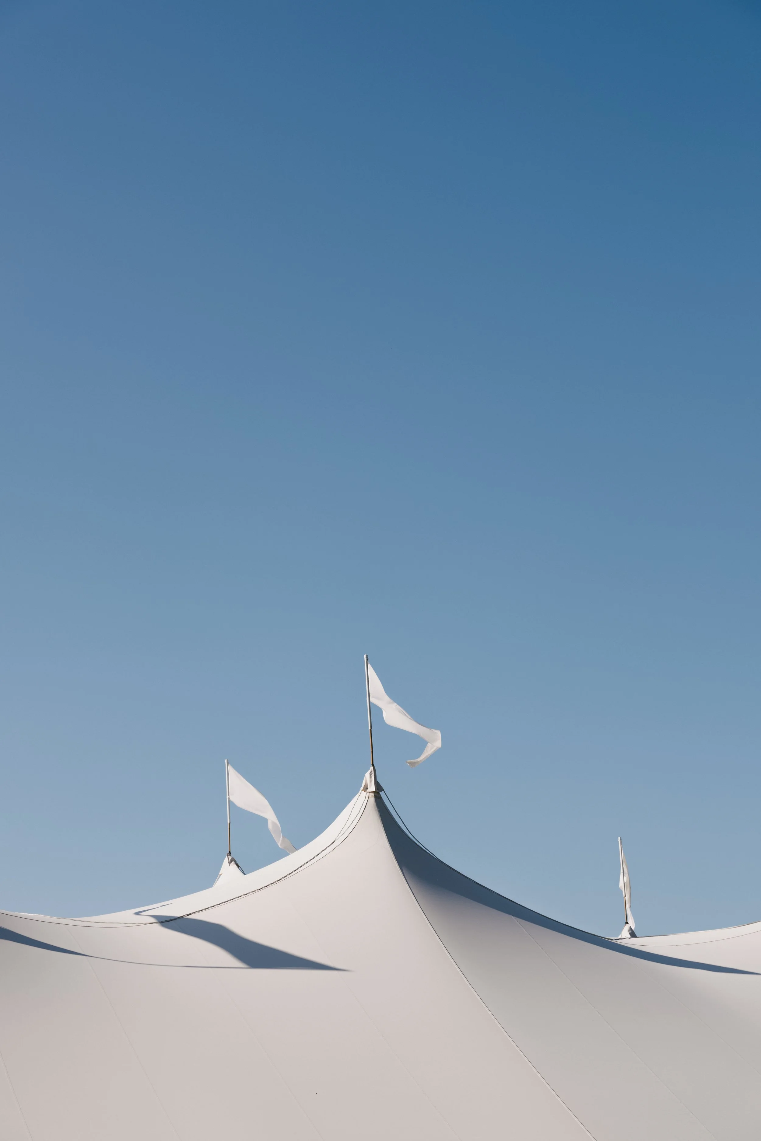 Close-up of a white fabric tent with peaked roof and small flags on top, set against a clear blue sky.