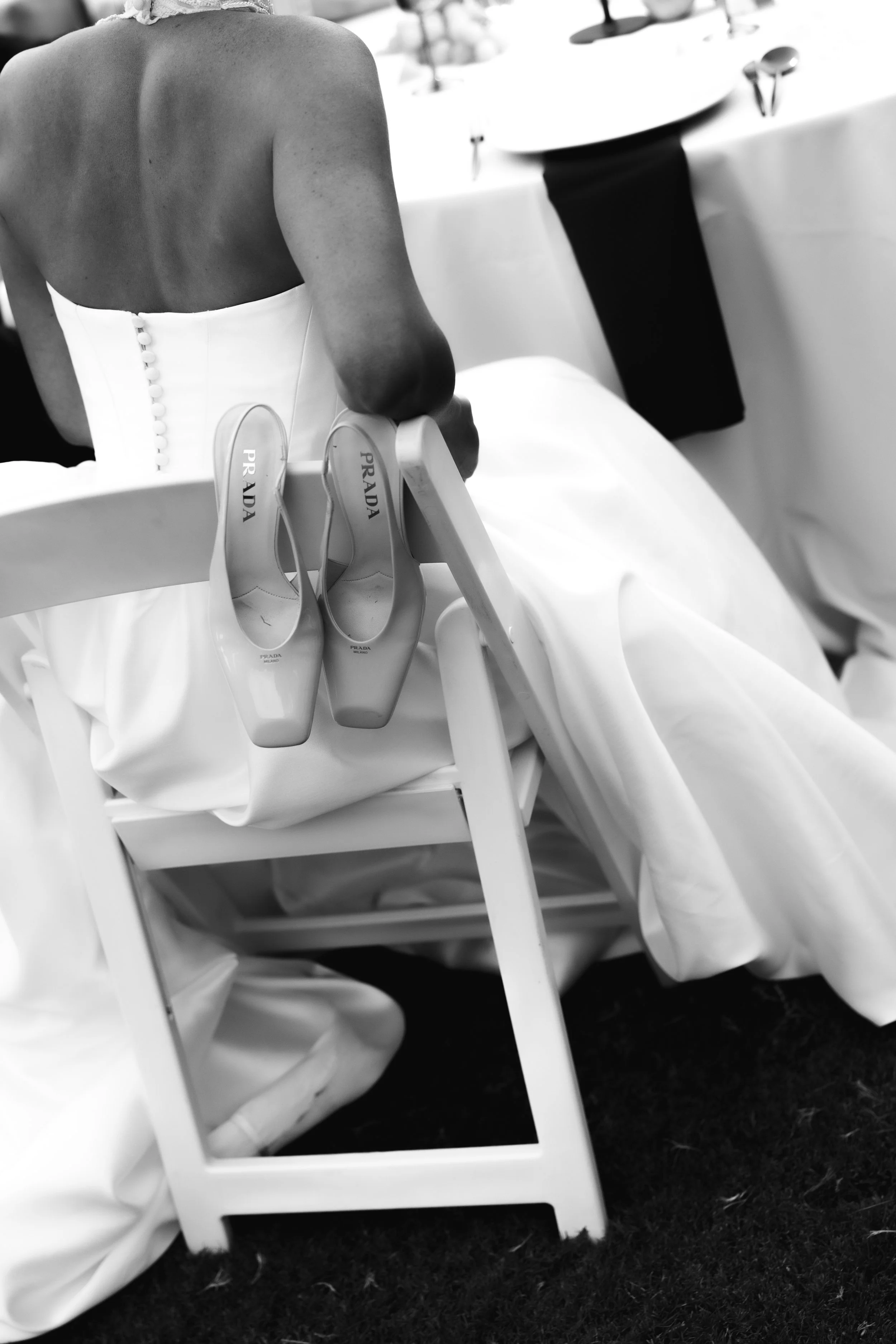 A woman in a white strapless dress sitting at a table with a pair of Prada high-heeled shoes hanging on the back of a white chair. The scene appears to be at a formal event like a wedding or banquet.