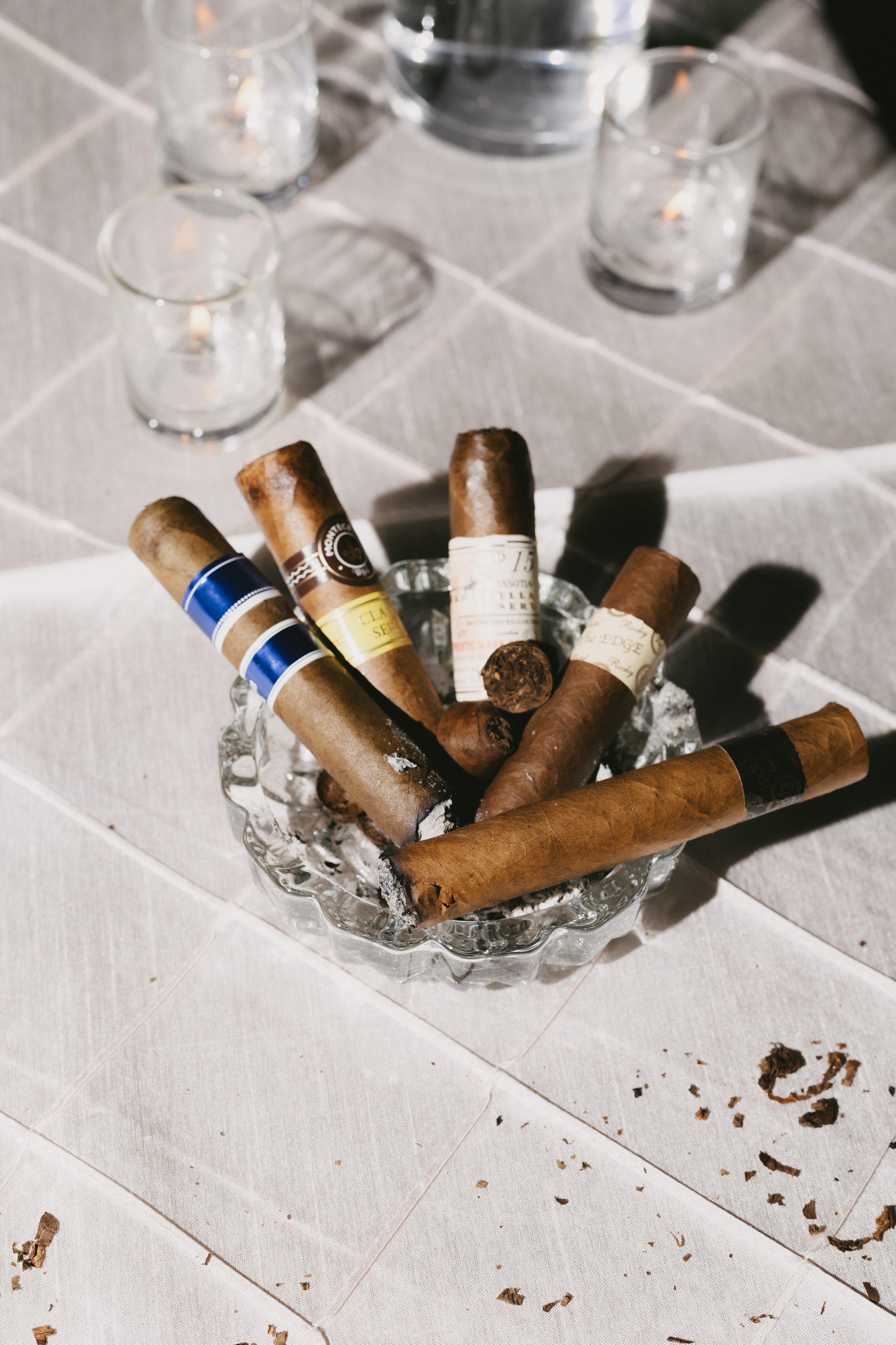 Assorted cigars in a glass bowl on a table with scattered cigar ashes and small debris.