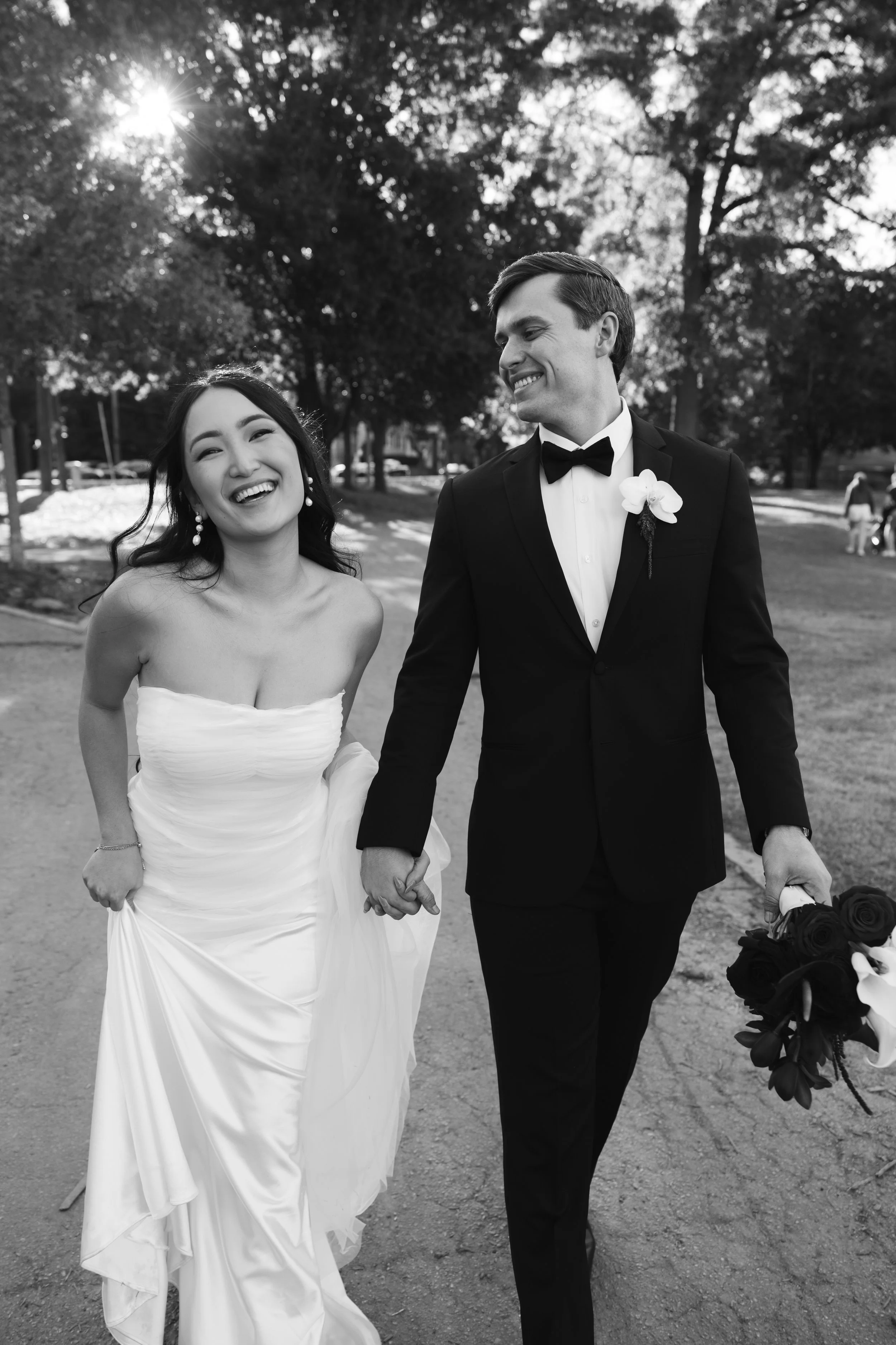 A bride and groom walking hand in hand outdoors, smiling, with trees and sunlight in the background.