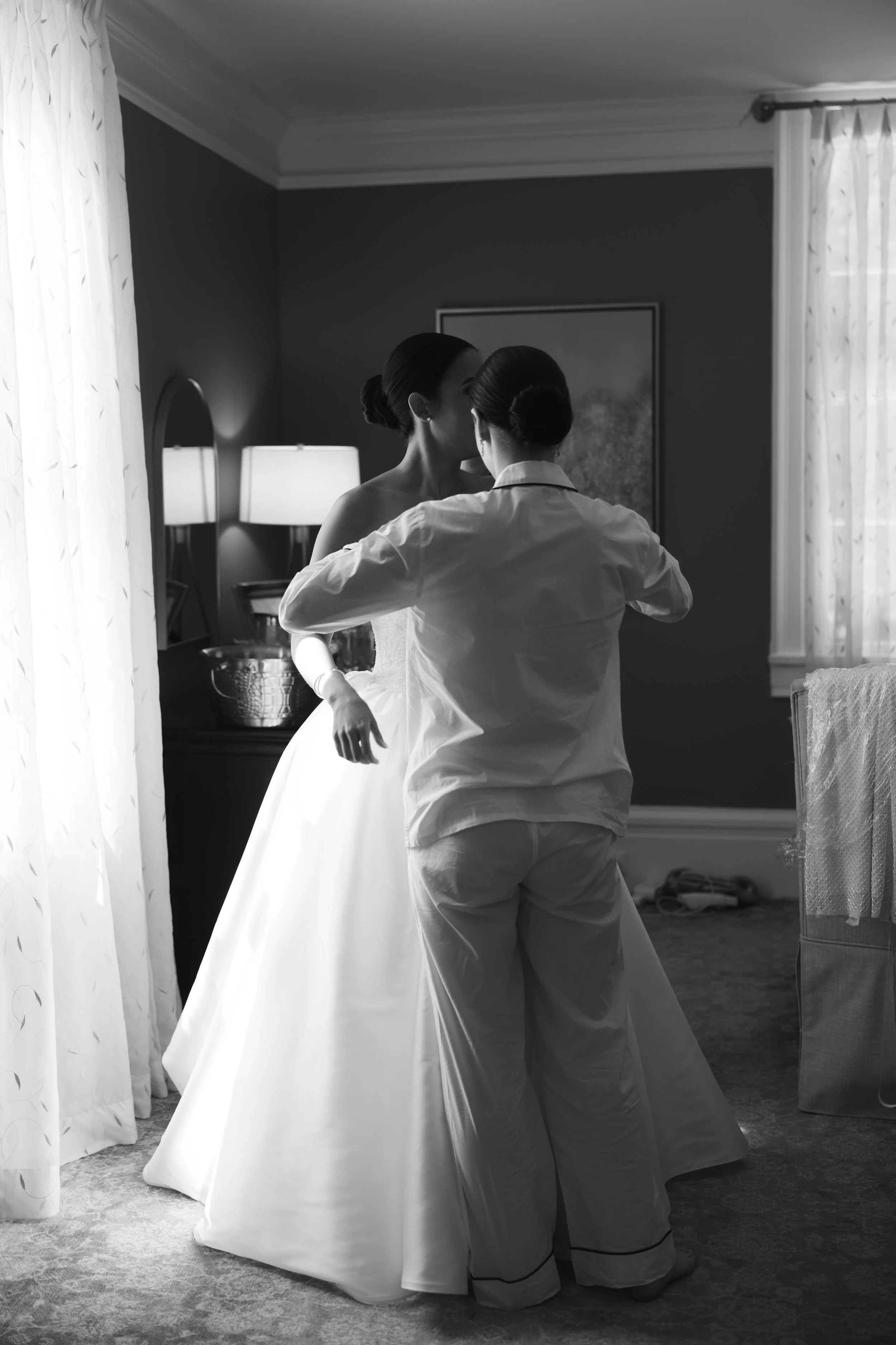 Black and white photo of a bride in a wedding dress being assisted with her dress by another woman in a room with a lamp, curtain, and framed artwork on the wall.