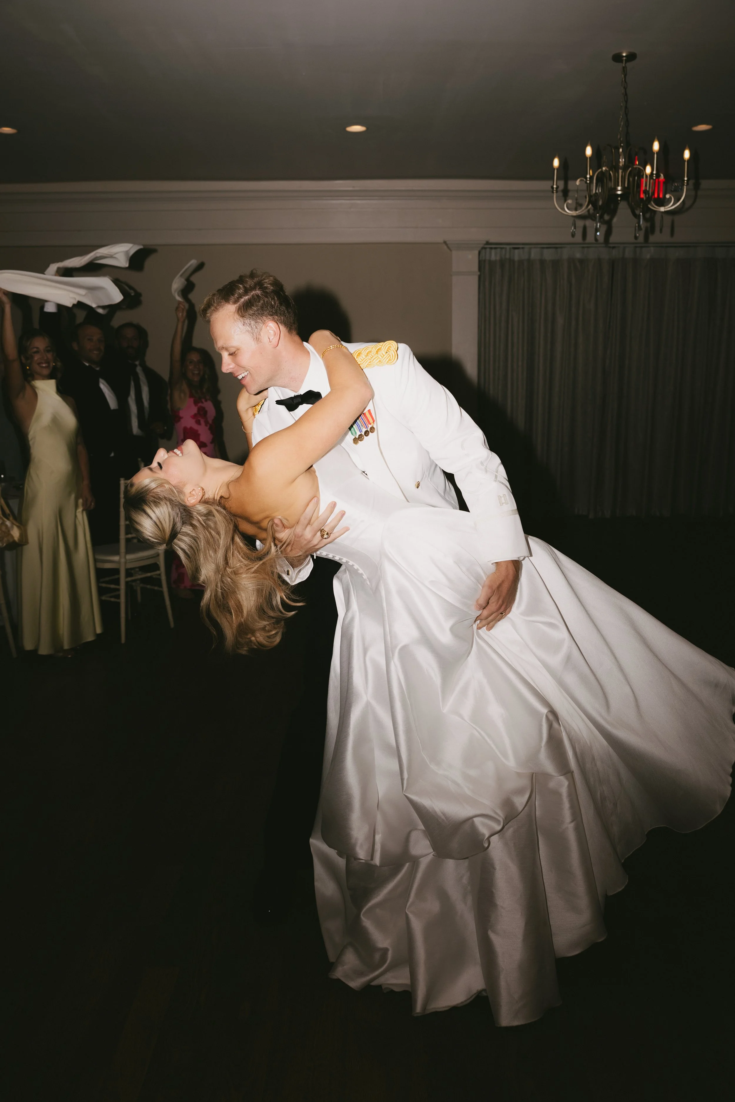 A couple dancing at a wedding reception, with the groom in a military uniform and the bride in a wedding gown, surrounded by friends and family.