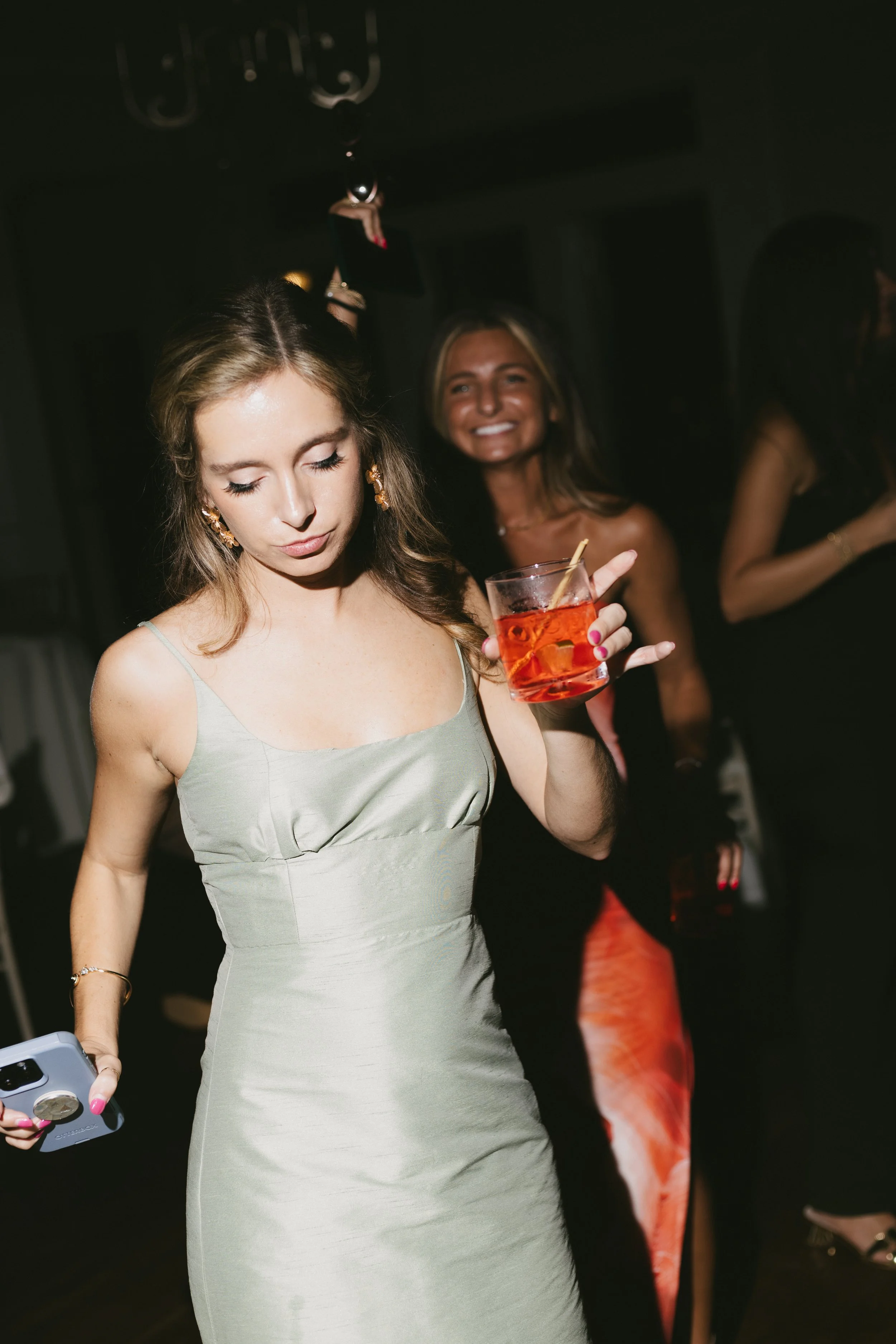 A young woman in a silver dress holding a cocktail at a party or nightclub, with other women in the background