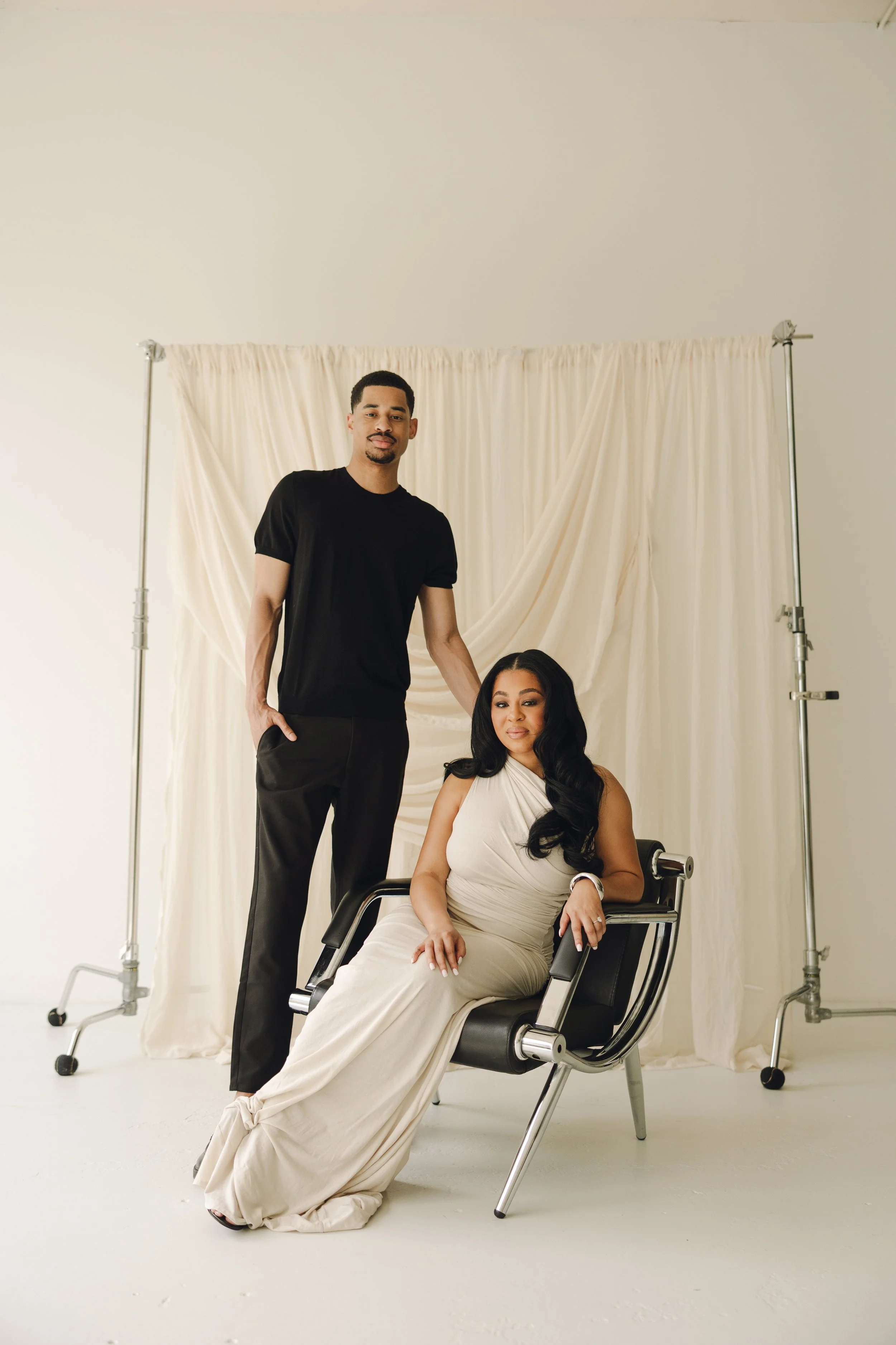 A man and a woman pose together in front of a light-colored backdrop. The woman is seated in a black chair, wearing a long, elegant cream-colored dress with black hair styled in loose waves. The man stands next to her, wearing a black t-shirt and bla