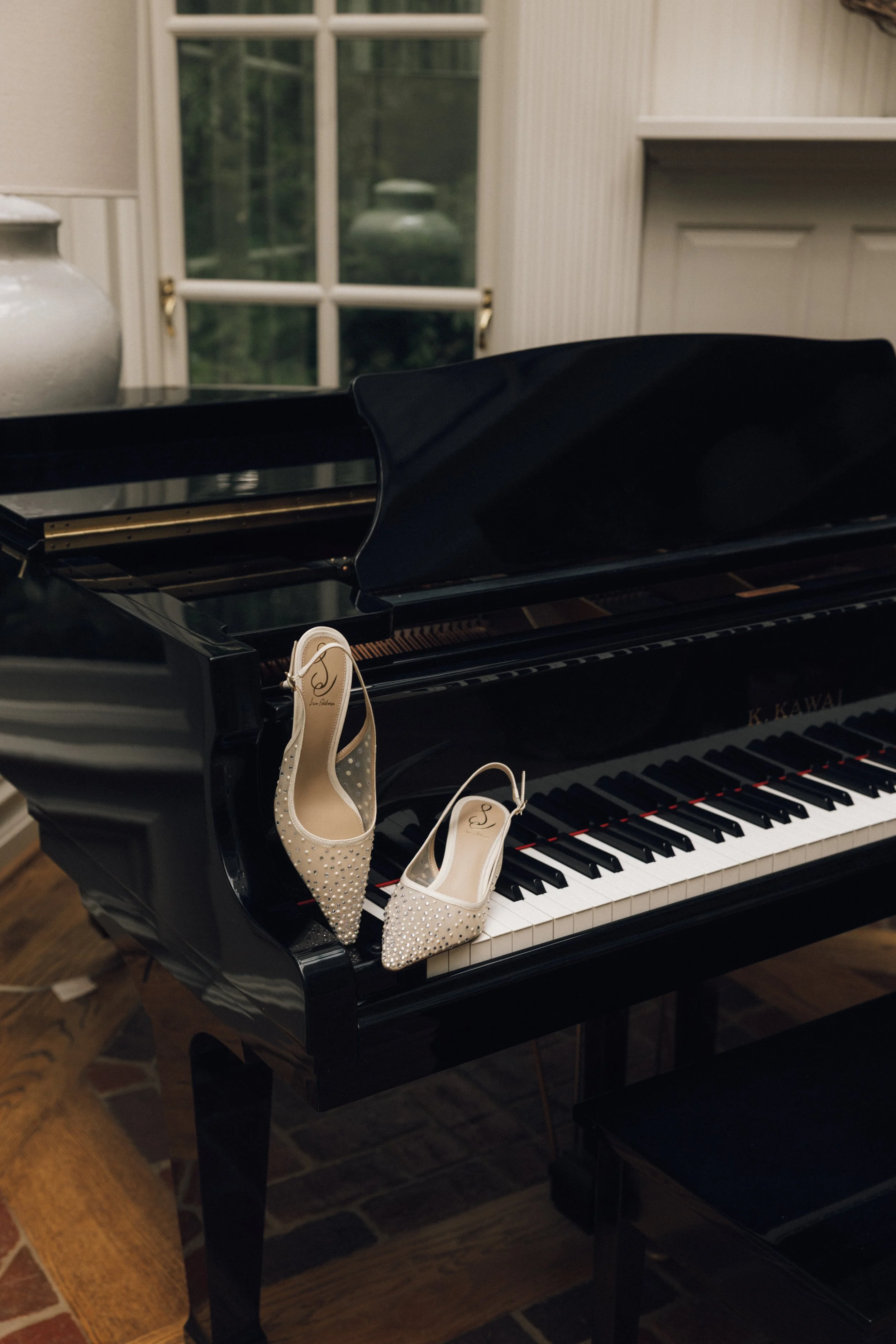 A pair of white, decorated high-heeled shoes with rhinestones sit on the keyboard of a black grand piano in a room with a window showing greenery outside.