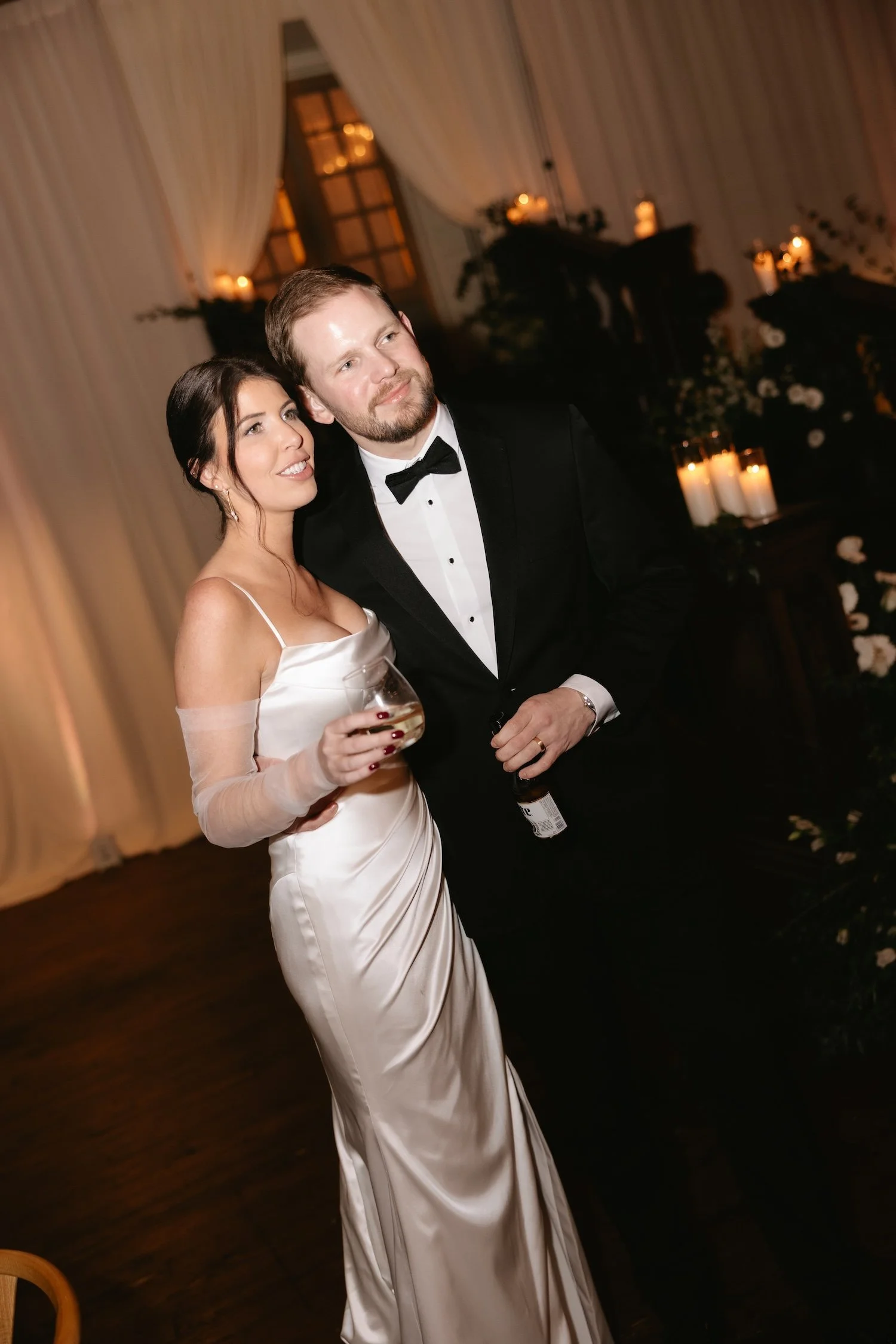 A bride in a white satin gown and a groom in a black tuxedo with a bow tie at a wedding reception, standing close together and smiling.