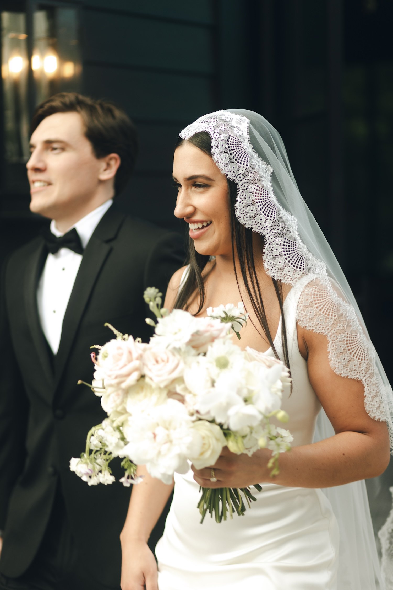 A bride holding a bouquet of white and pale pink flowers, wearing a white dress and lace-edged veil, smiling. A groom in a black tuxedo is standing beside her, slightly out of focus.