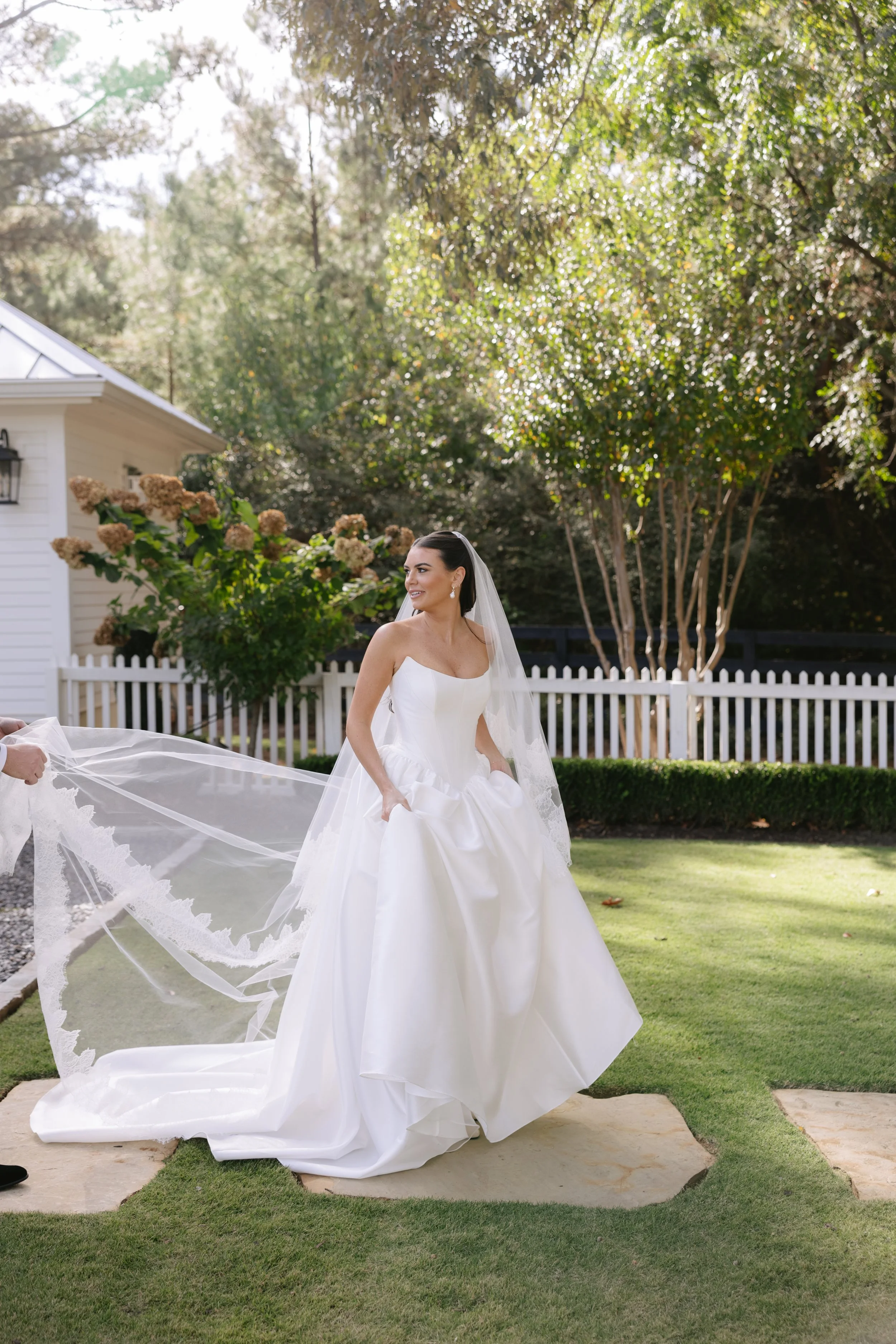 A bride in a white wedding dress and veil standing on a stone pathway in a garden, smiling and holding her dress.