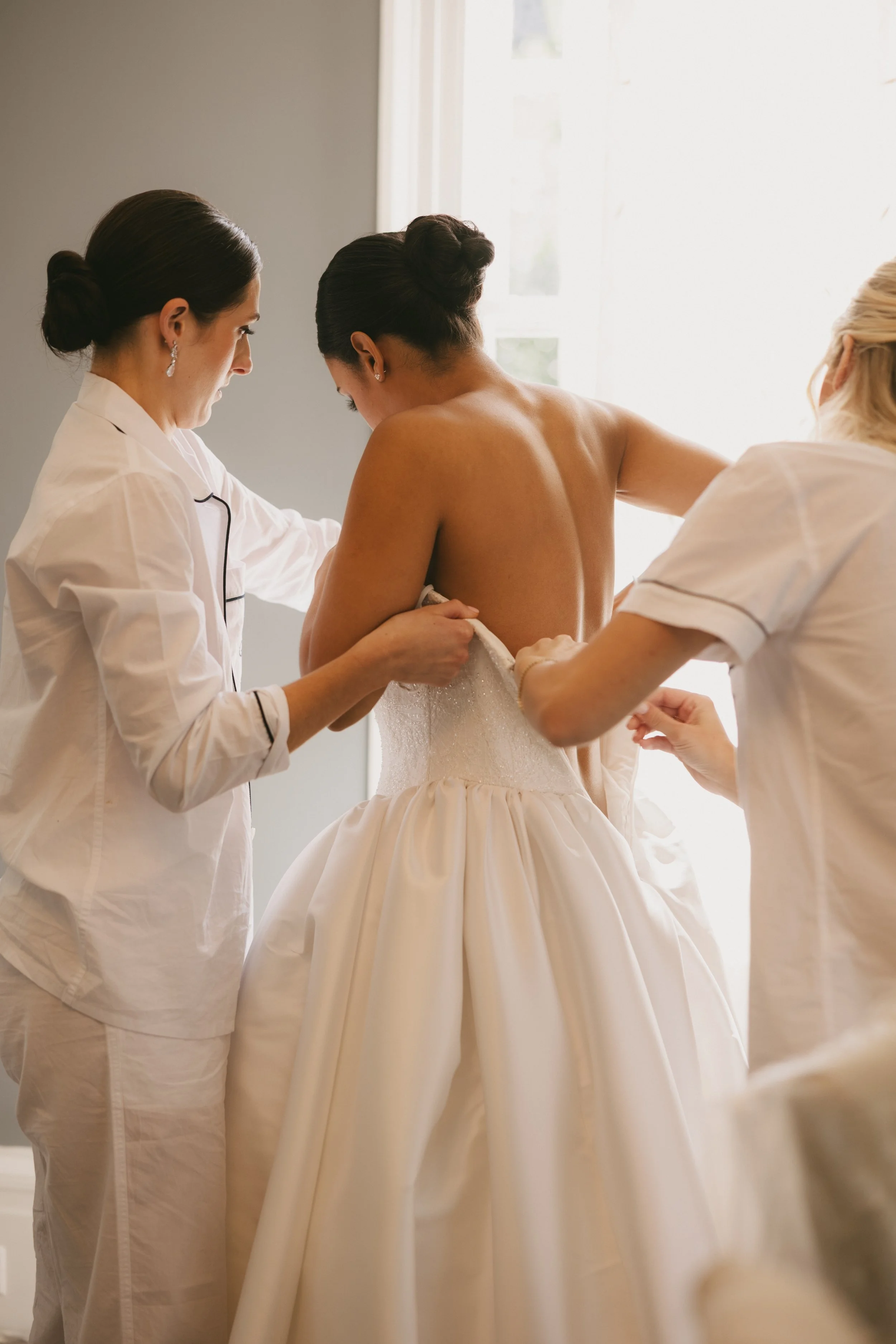 Three women assist a bride in putting on her wedding dress by a window in a well-lit room.