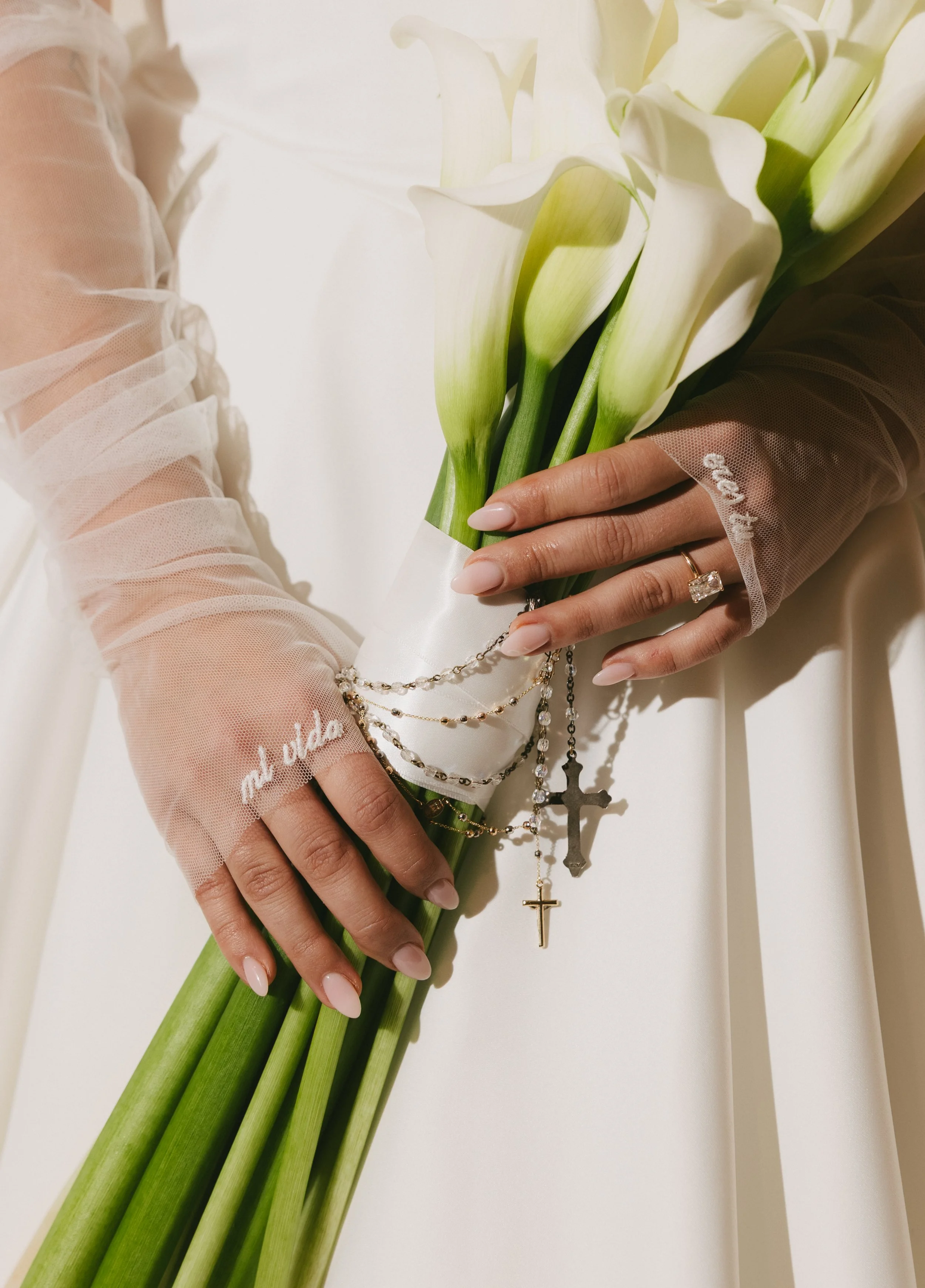 Person holding a bouquet of white calla lilies with jewelry and a wedding ring, wearing sheer, embroidered sleeves.