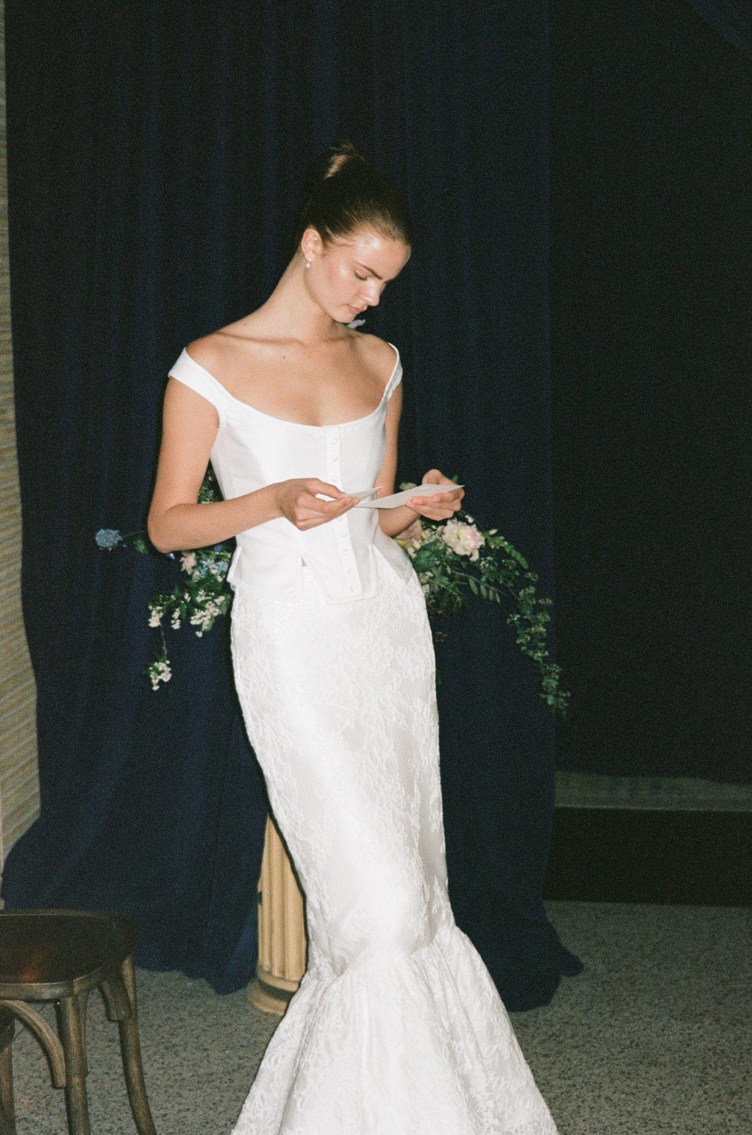 A woman in a white wedding dress reading a note or speech at her wedding ceremony, standing in front of dark curtains and floral arrangements.