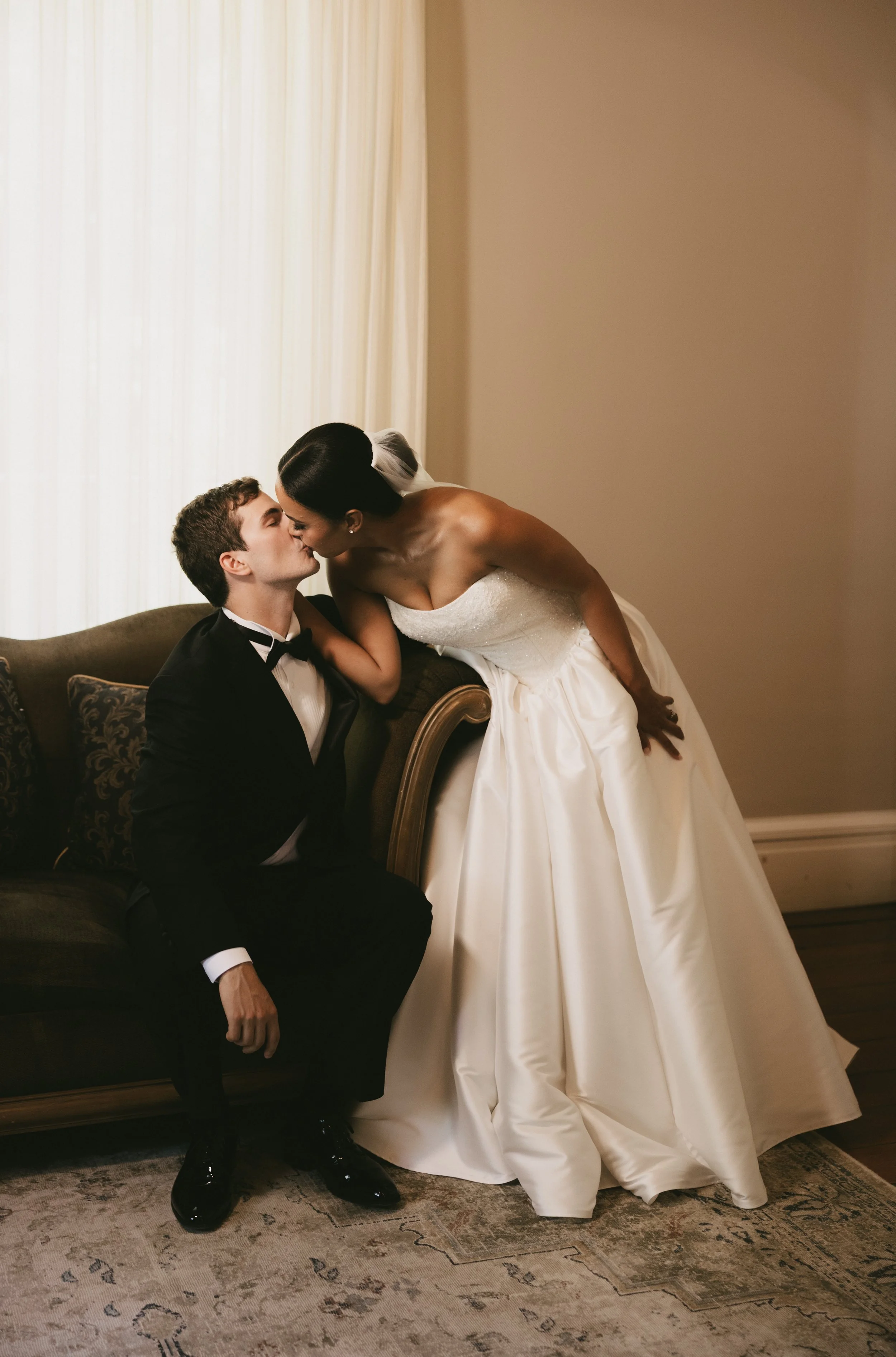 A bride and groom sharing a kiss indoors, with the bride leaning over a couch to kiss the groom, who is seated on the edge of the couch. The bride is wearing a strapless white wedding dress, and the groom is in a black tuxedo with a bow tie.