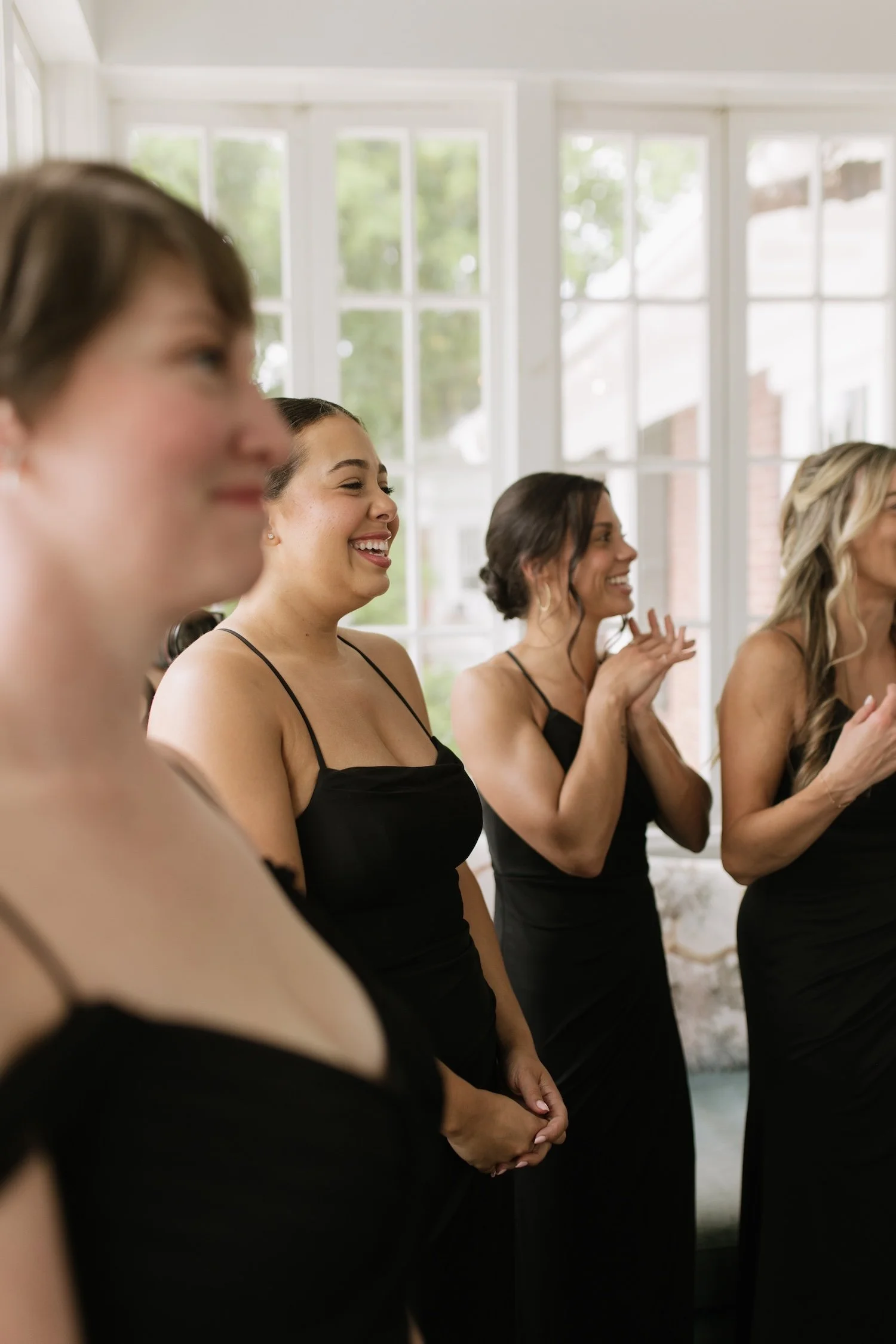 Four women dressed in black dresses standing indoors near large windows, smiling and clapping, possibly at a special event or celebration.