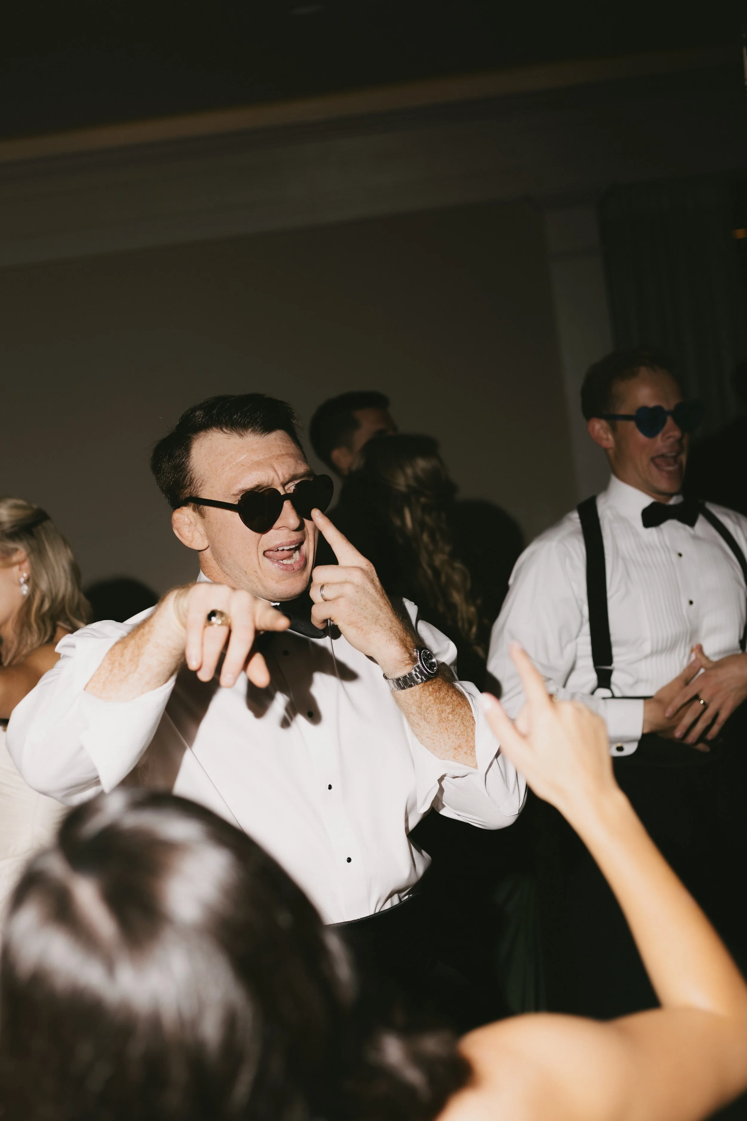 People dancing at a party, wearing tuxedos, with some wearing sunglasses, in a low-light setting.