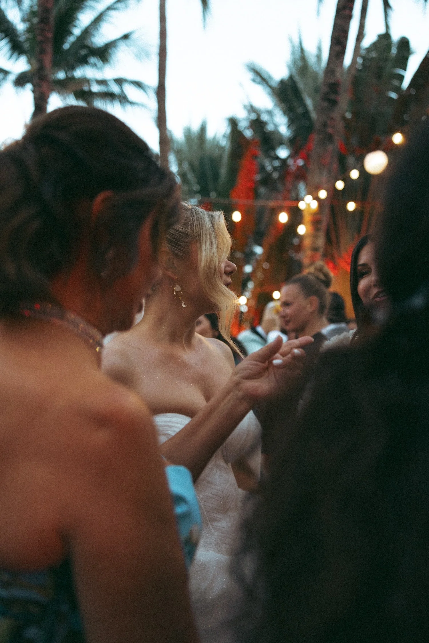 Group of women talking and smiling outdoors, with palm trees and string lights in the background during an evening event.