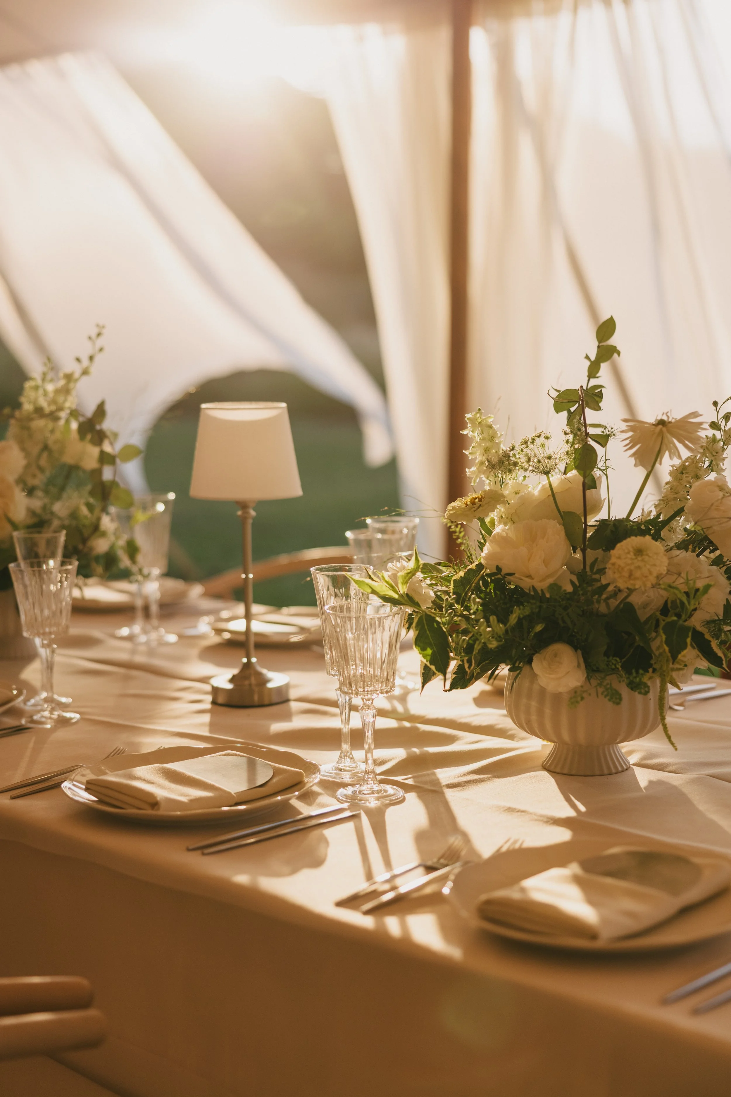 A beautifully set dining table inside with sunlight streaming through draped curtains, decorated with a large floral centerpiece, glassware, and a table lamp.
