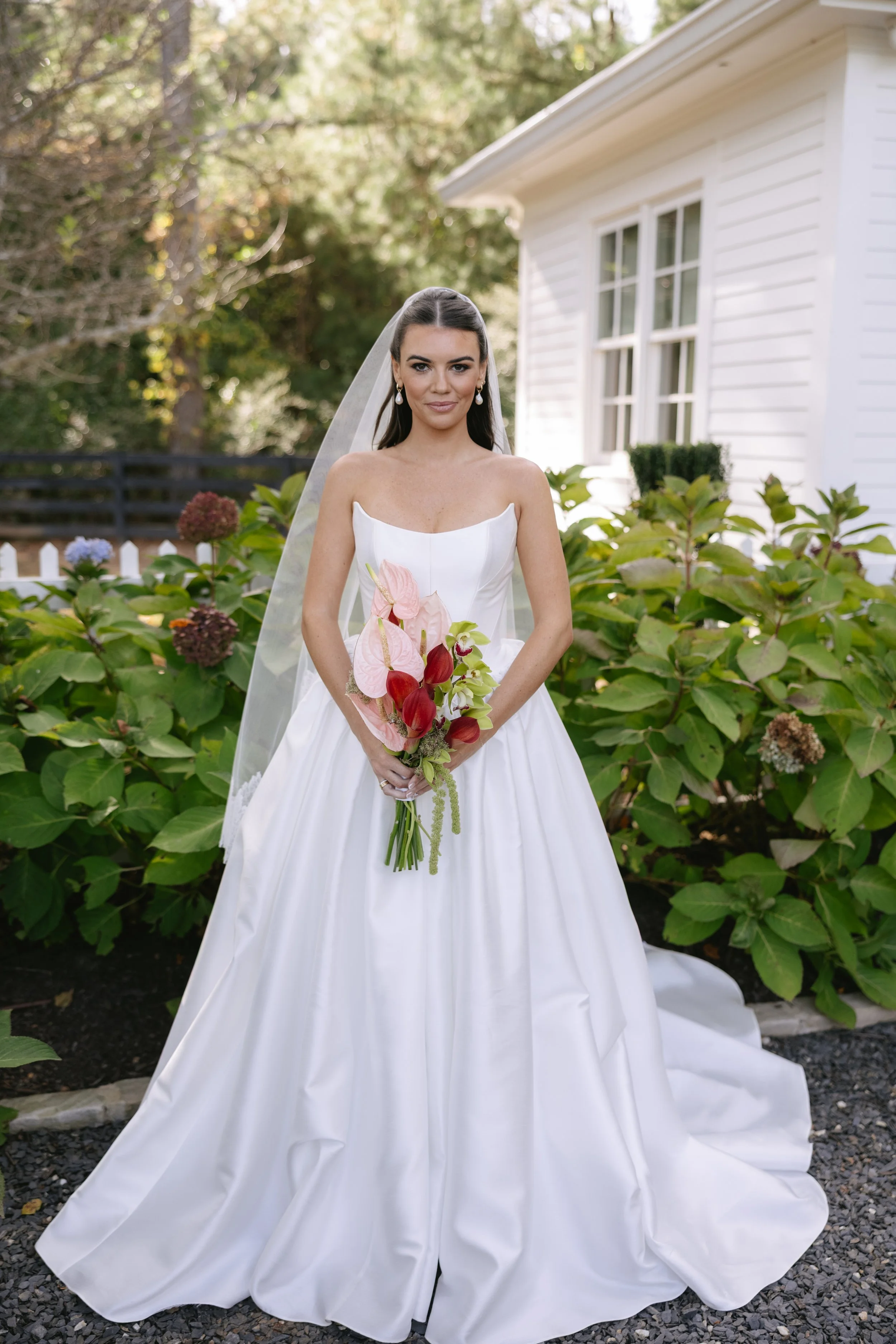 A bride in a strapless white wedding gown holding a bouquet of pink and red flowers, standing outdoors in front of green bushes and a white house with a window.