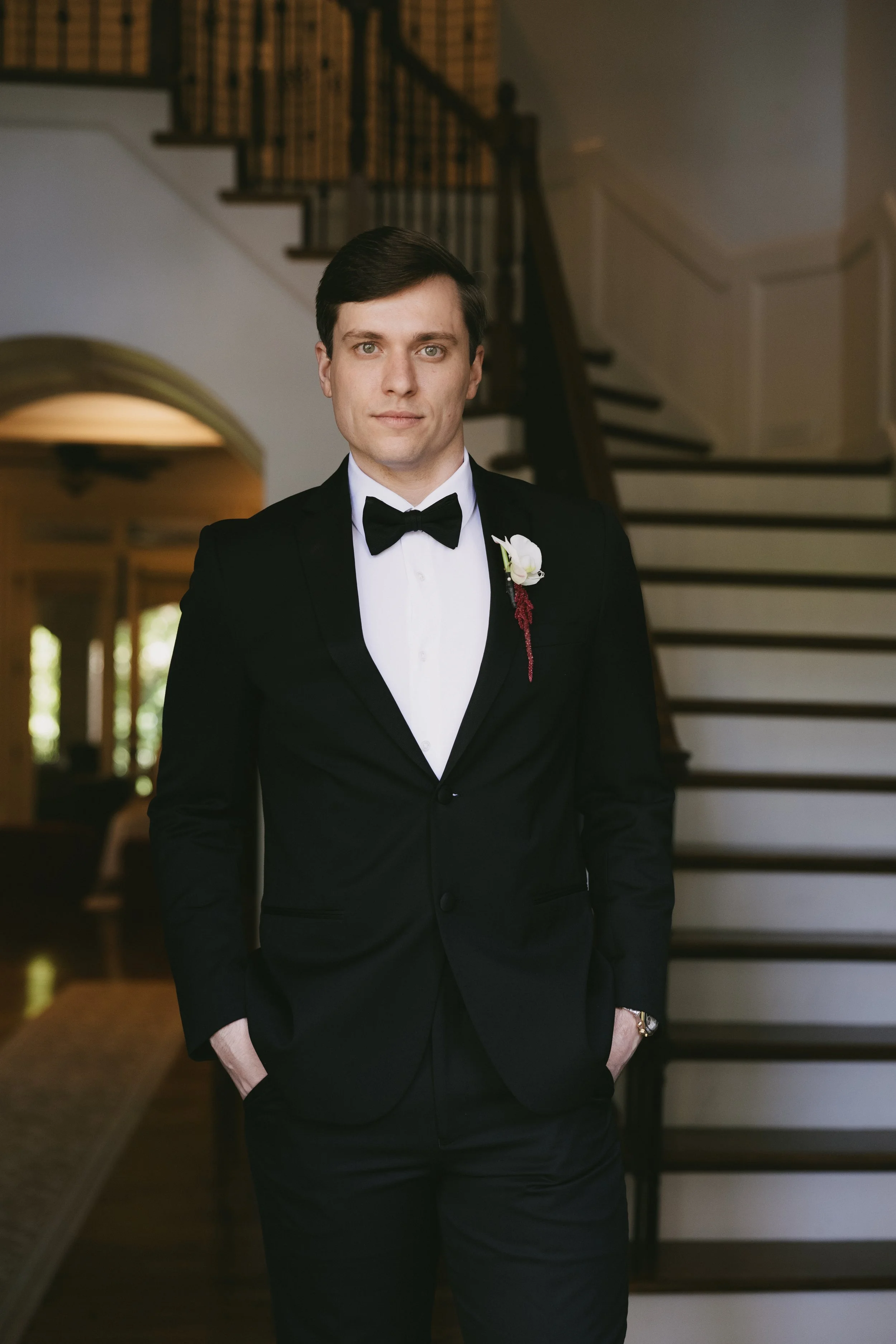 Man in a black tuxedo with a bow tie, standing indoors near stairs, with his hands in his pockets.