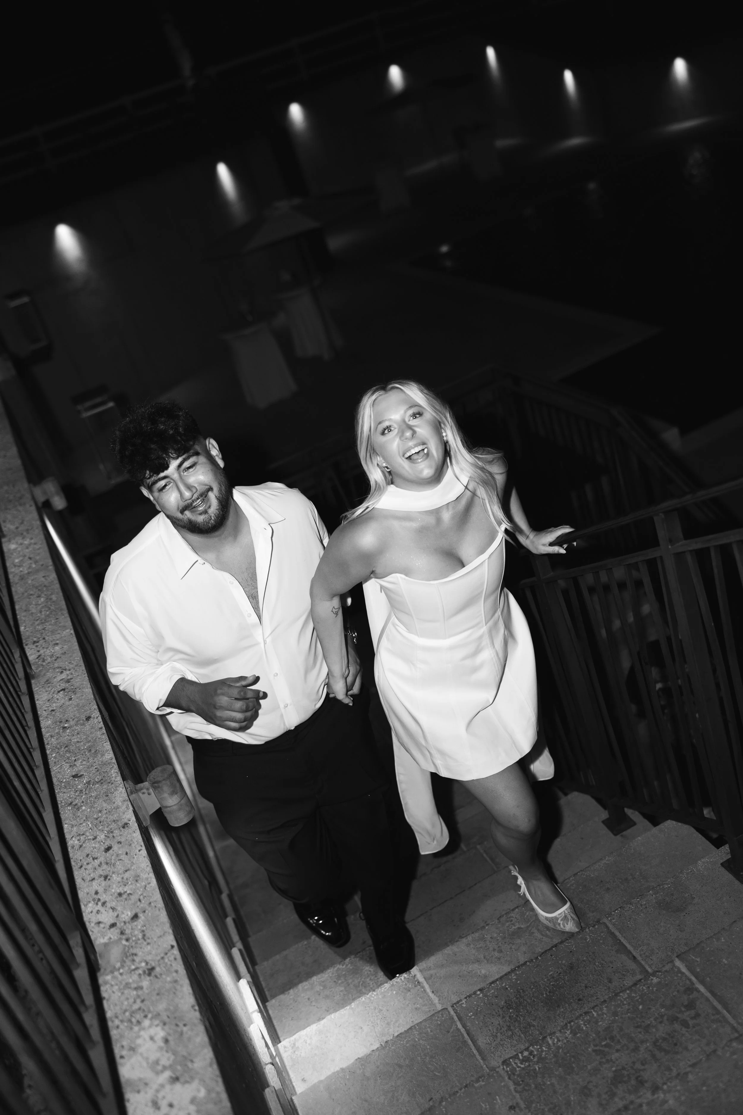 A black and white photo of a man and woman ascending stairs, the woman is smiling and wearing a white dress, and the man is smiling and wearing a white shirt and dark pants, at an indoor venue with ceiling lights.