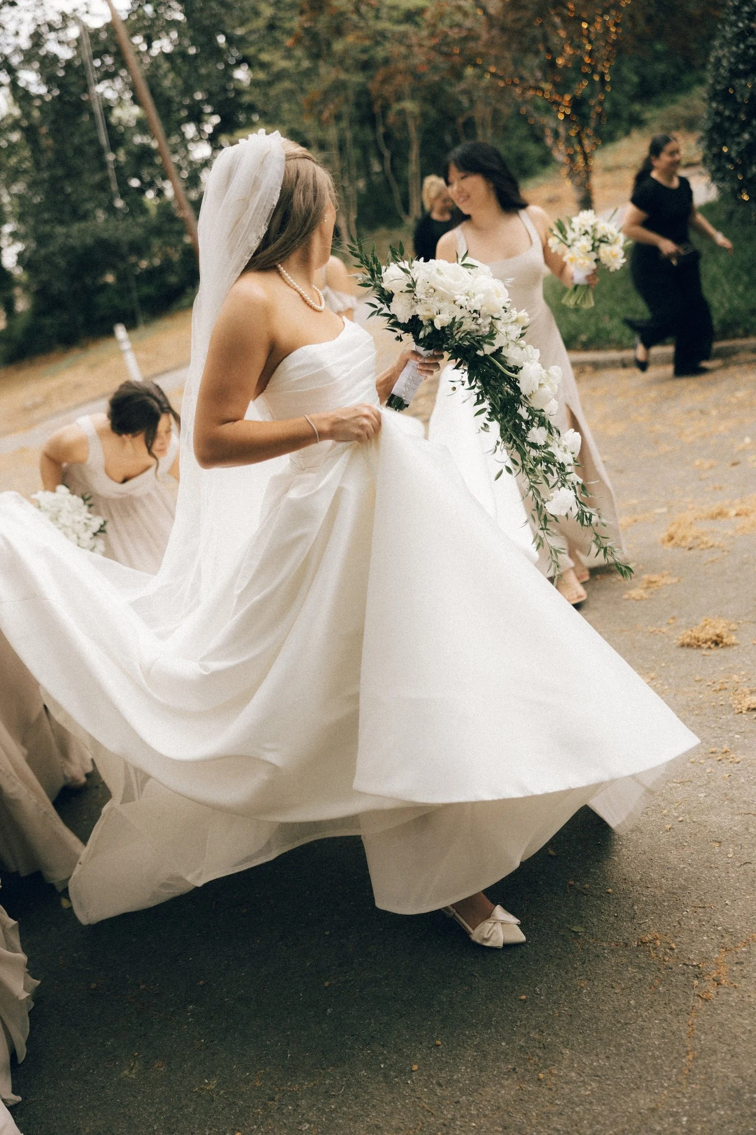 A bride in a white wedding dress and veil holding a large bouquet of white flowers and greenery, surrounded by bridesmaids in light-colored dresses, outdoors with trees and string lights in the background.