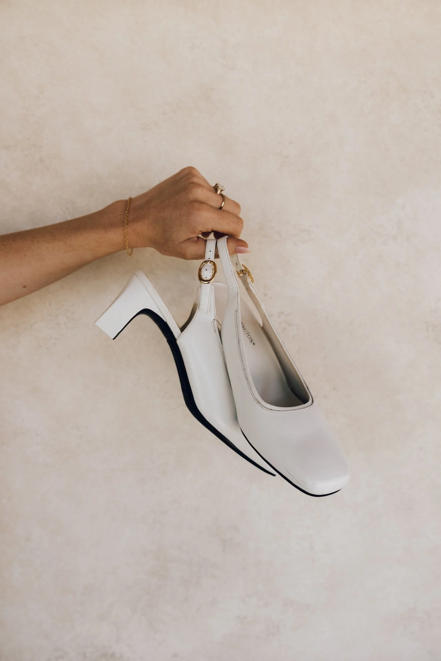 Hand holding a white high-heeled shoe with black sole against a beige wall.