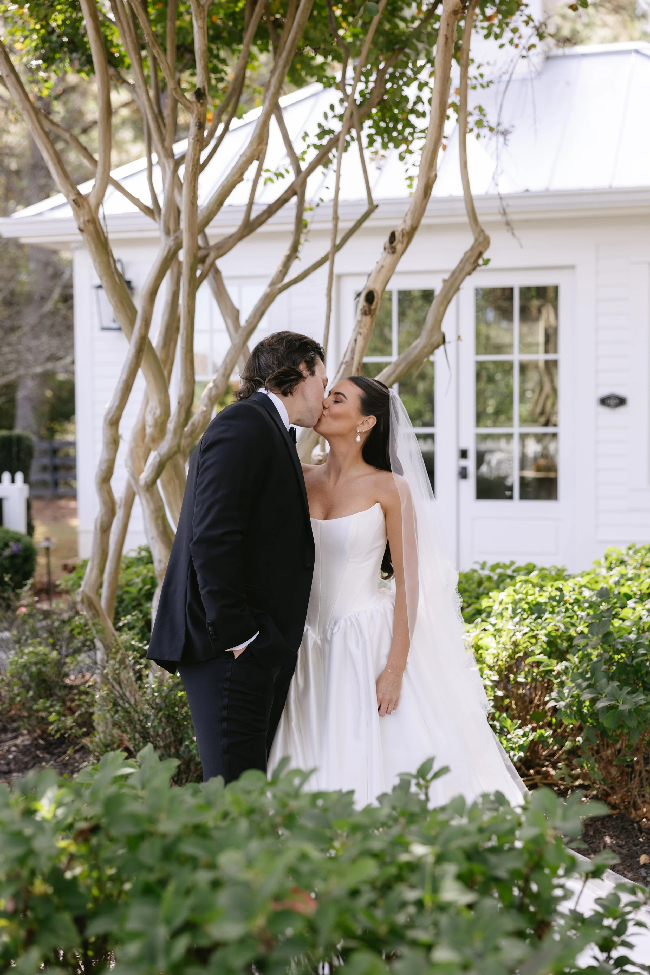 A newlywed couple sharing a kiss outdoors amidst greenery and a tree, with a white house in the background.