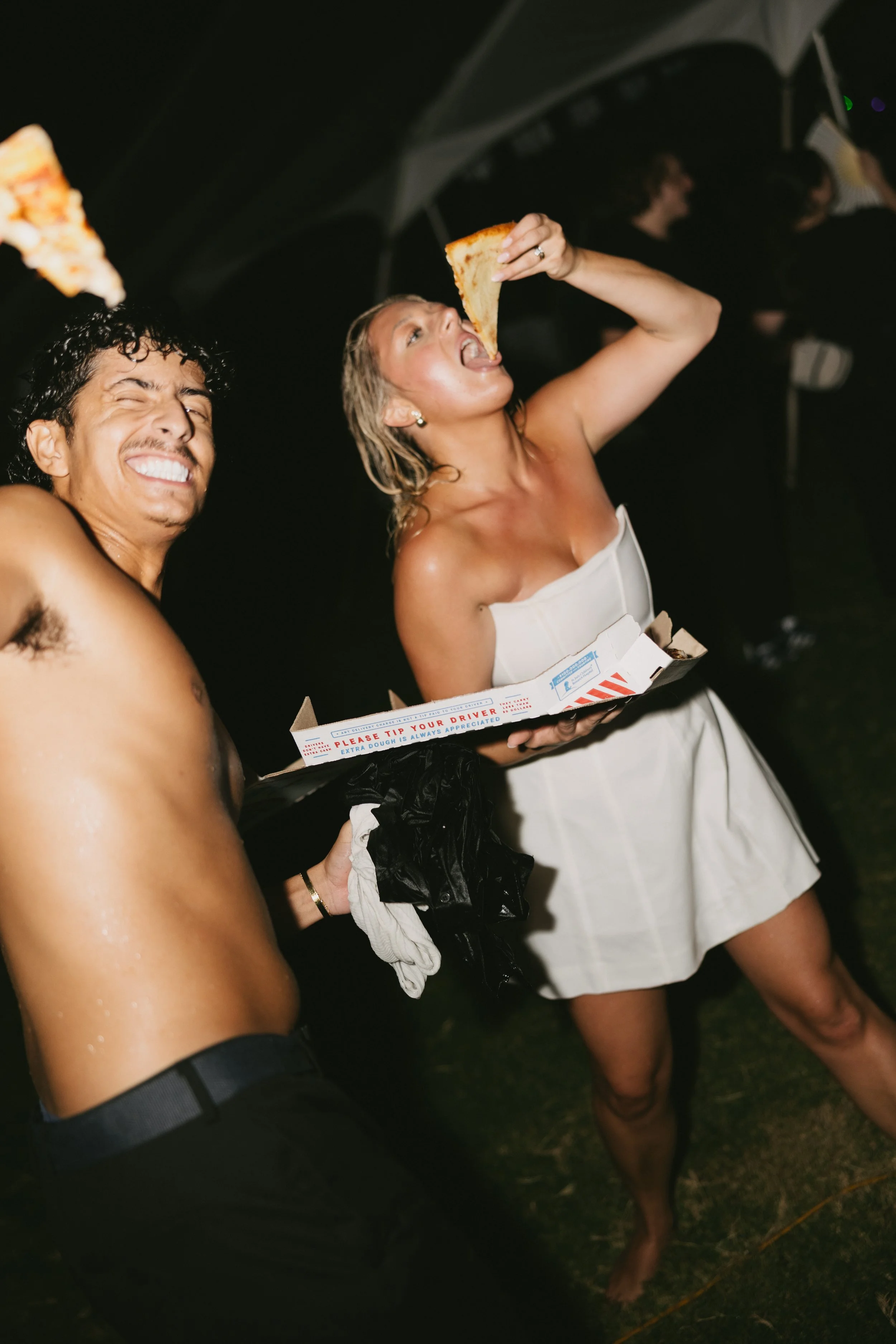 A shirtless man and a woman in a white dress are enjoying pizza at a party.