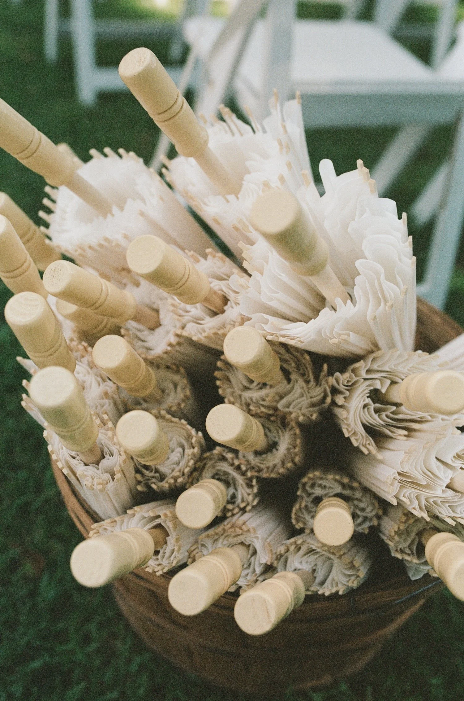 A basket filled with bundled white paper umbrellas with wooden handles, set outdoors on green grass.