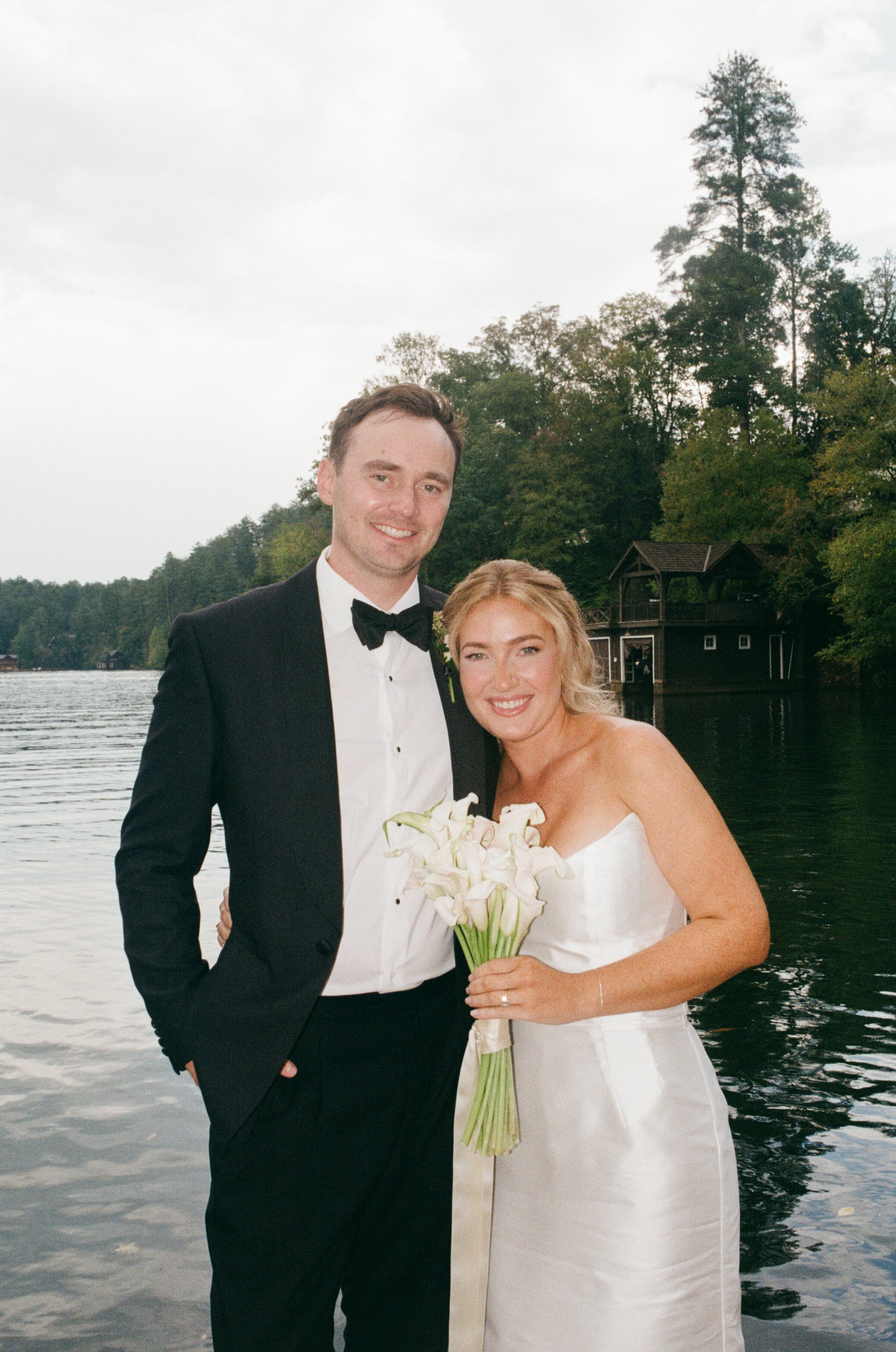 A newlywed couple standing by a lake, smiling at the camera. The groom is wearing a black tuxedo and the bride is holding a bouquet of white calla lilies.