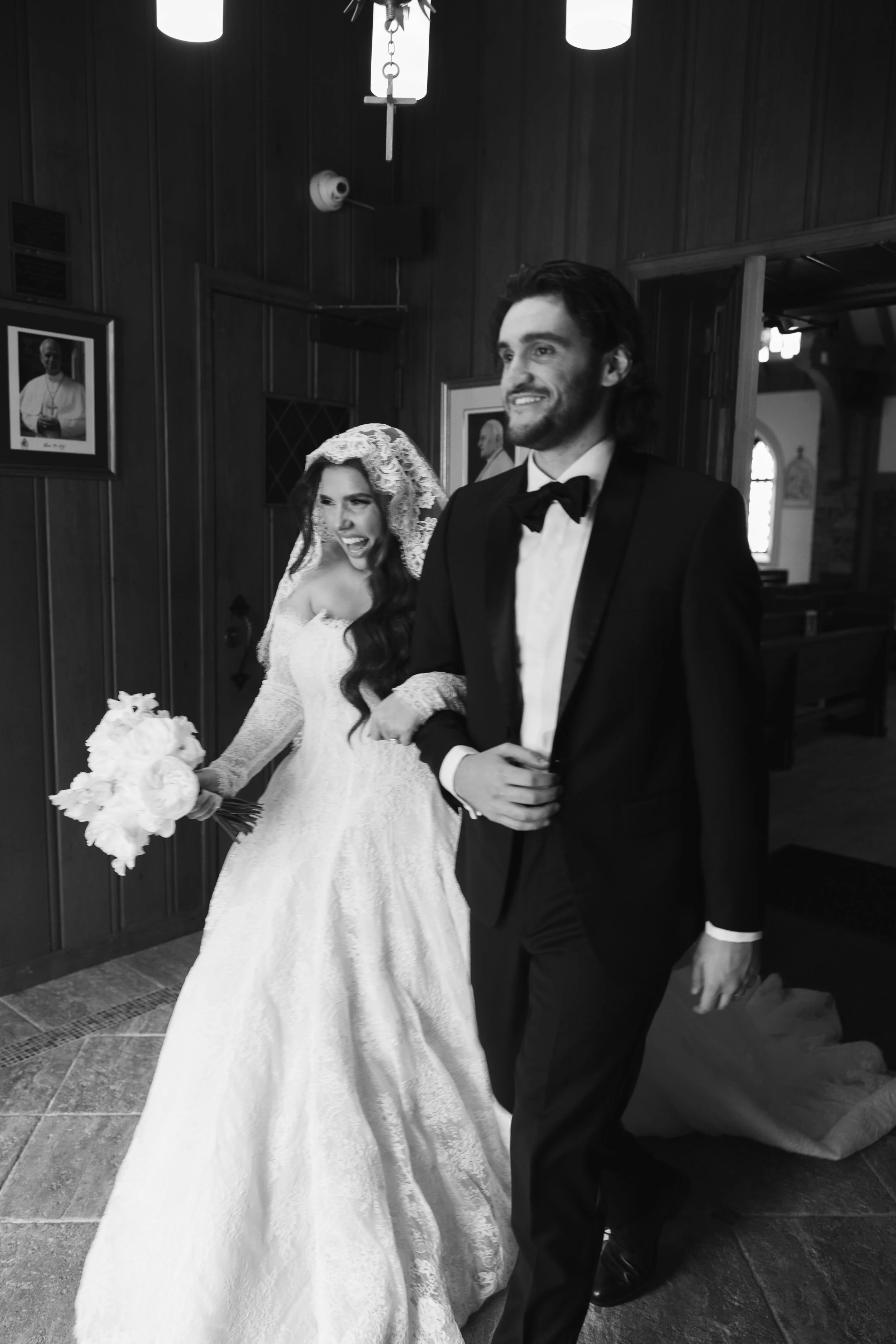 Black and white photo of a bride and groom walking inside a church. The bride is wearing a wedding dress and holding a bouquet, and the groom is in a tuxedo. They are both smiling happily.