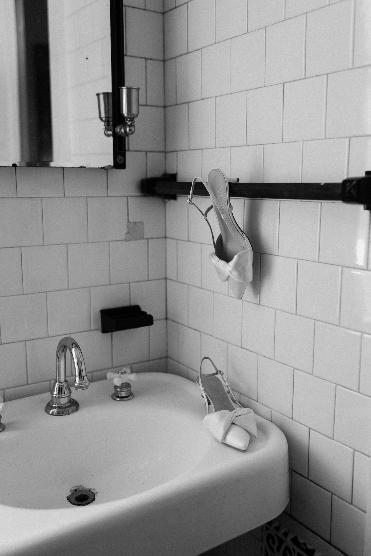 Black and white photo of a bathroom sink with a mirror, two high heels with bows hanging on a rail, and a mask hanging from one of the shoes.
