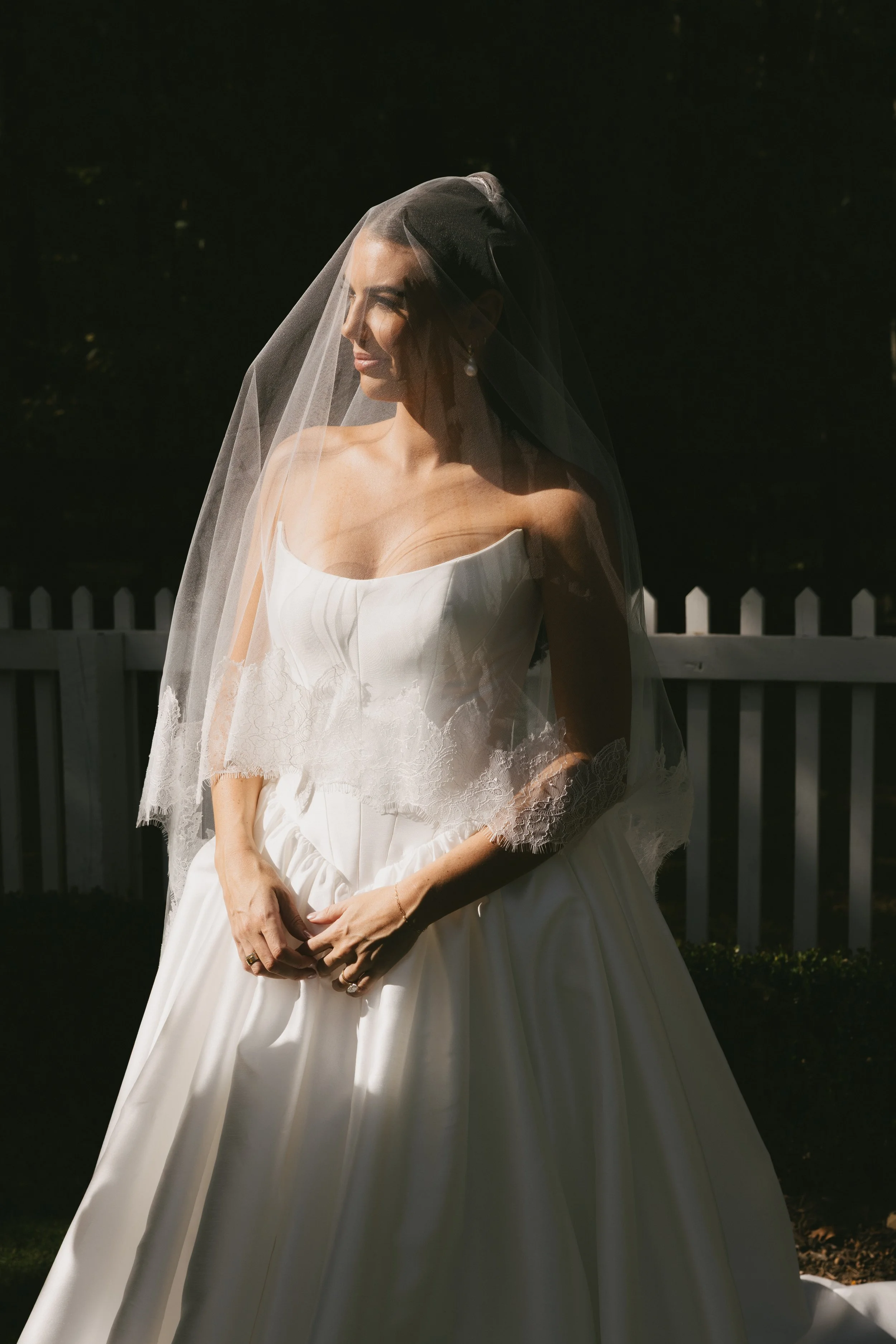 A bride in a white wedding dress with lace details, standing outdoors with a white picket fence behind her, gently smiling and looking to her left.