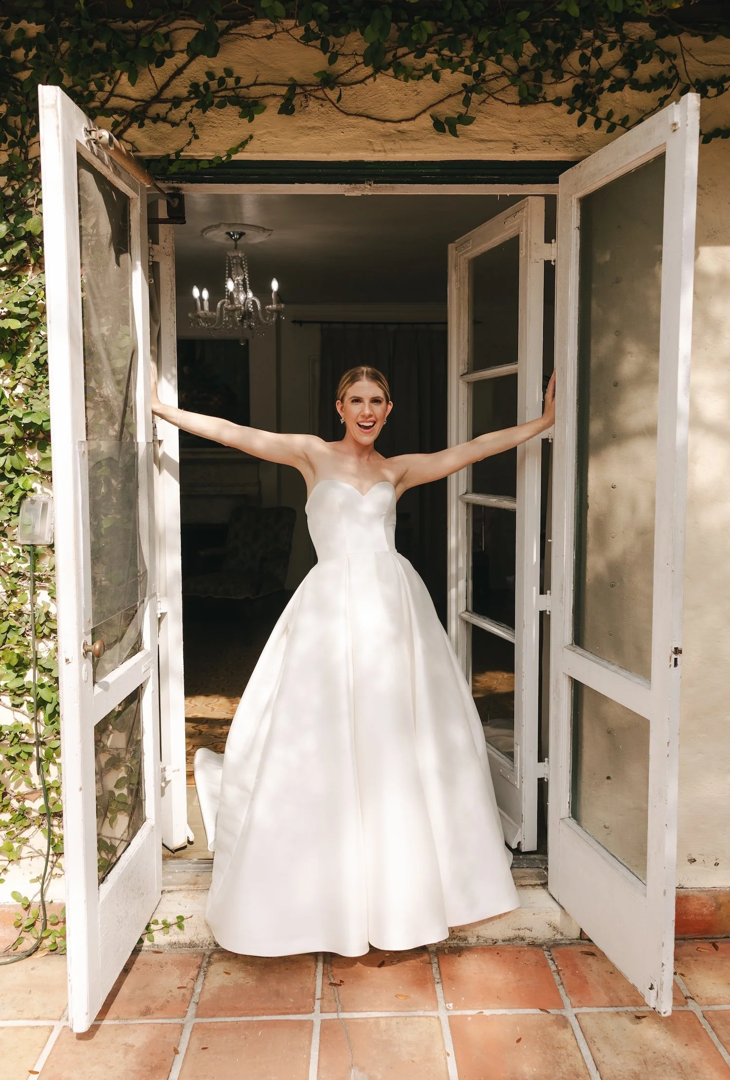 A woman in a white wedding dress standing in the open doorway of a house, smiling with arms outstretched.