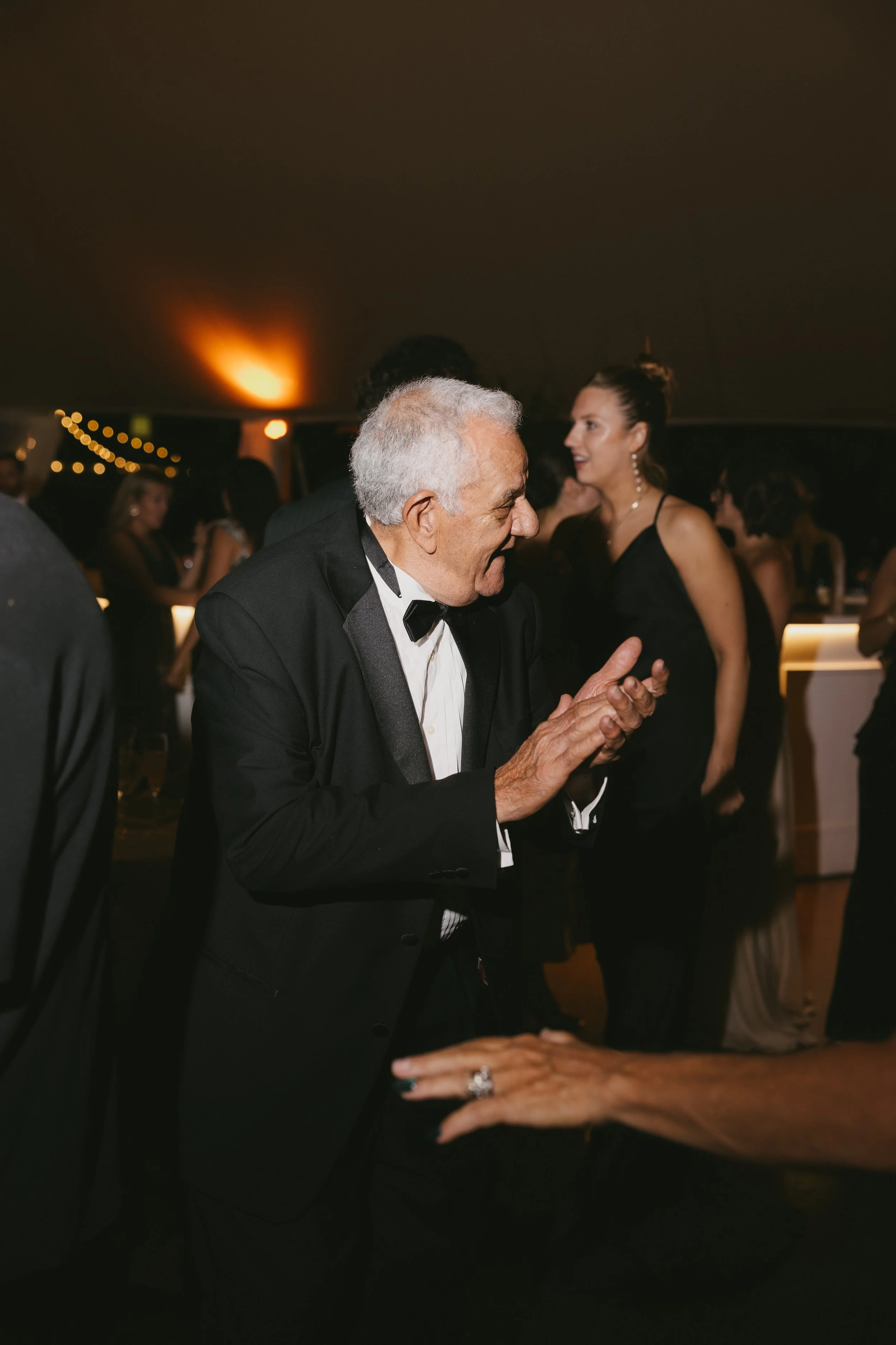 An elderly man in a tuxedo dancing and clapping at a formal event, with a woman in a black dress and other guests in the background.