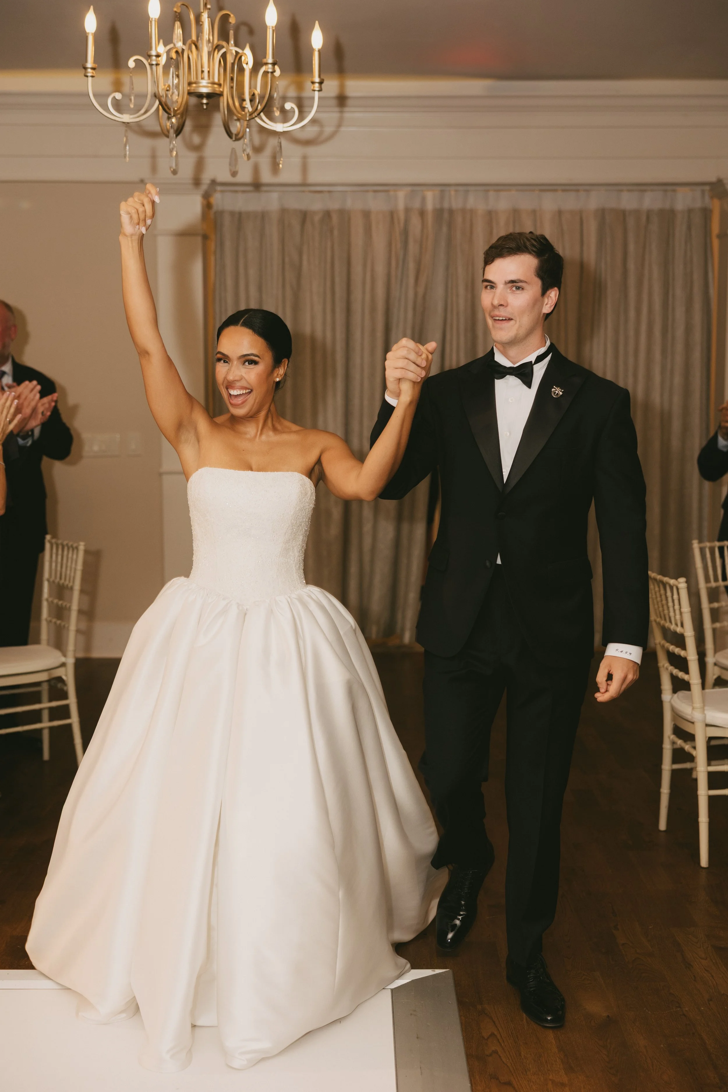A newlywed couple, the bride in a white strapless wedding gown and the groom in a black tuxedo, holding hands and celebrating at their wedding reception. The bride is smiling and raising her arm while the groom looks pleased. Guests are clapping in t