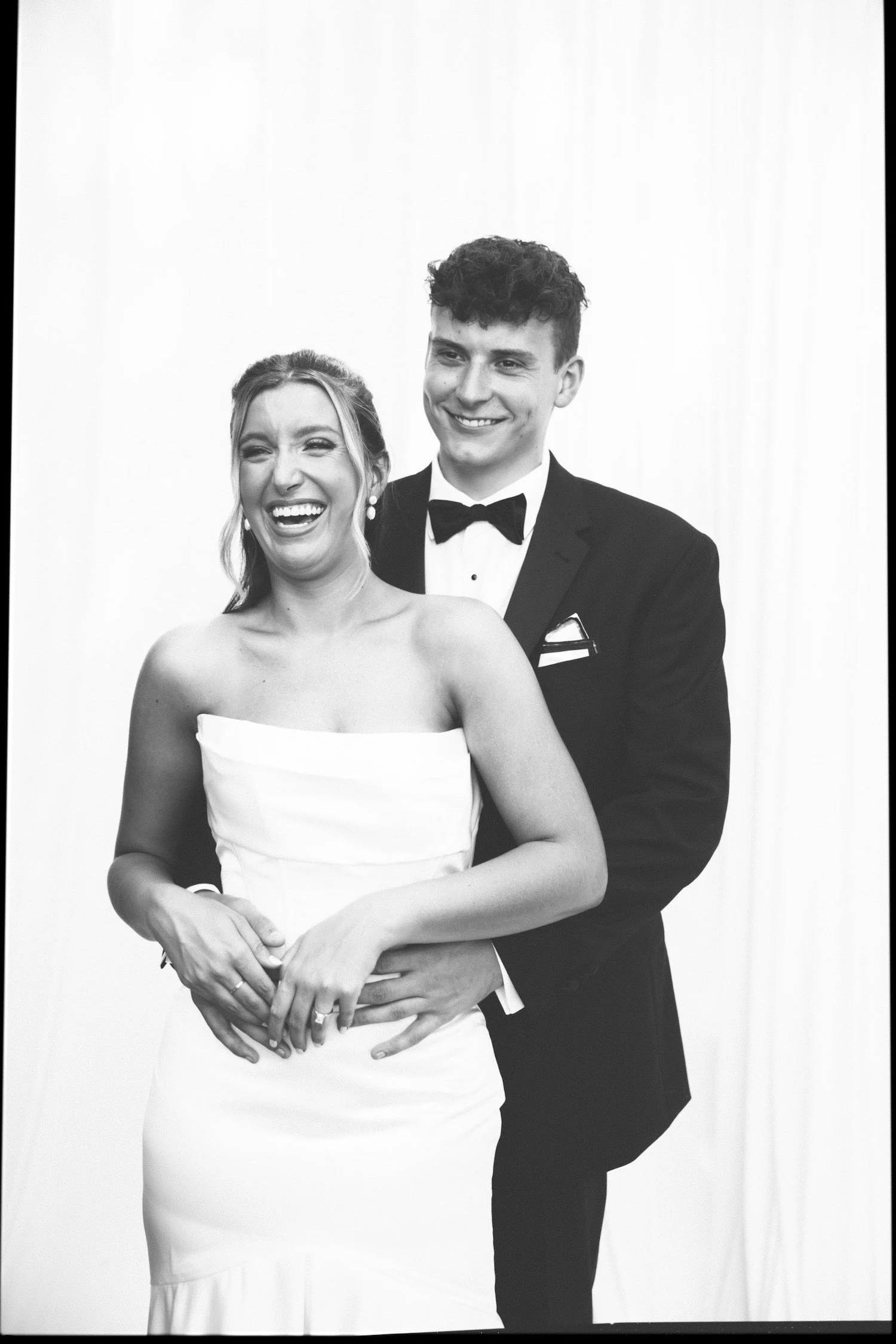 A joyful woman in a strapless white wedding dress and a man in a tuxedo stand close together, smiling and laughing.