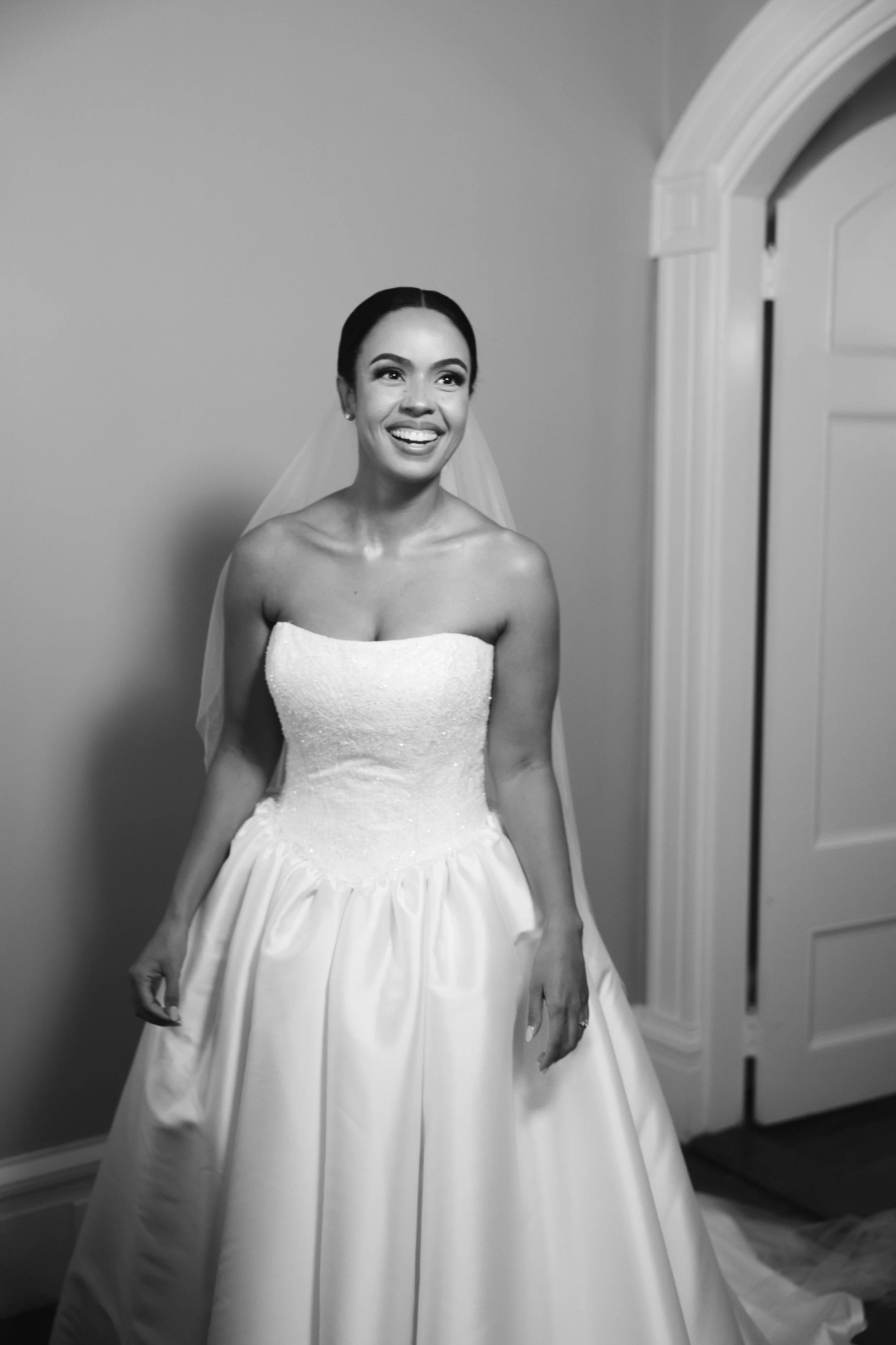 A smiling bride in a wedding dress standing indoors near a door.