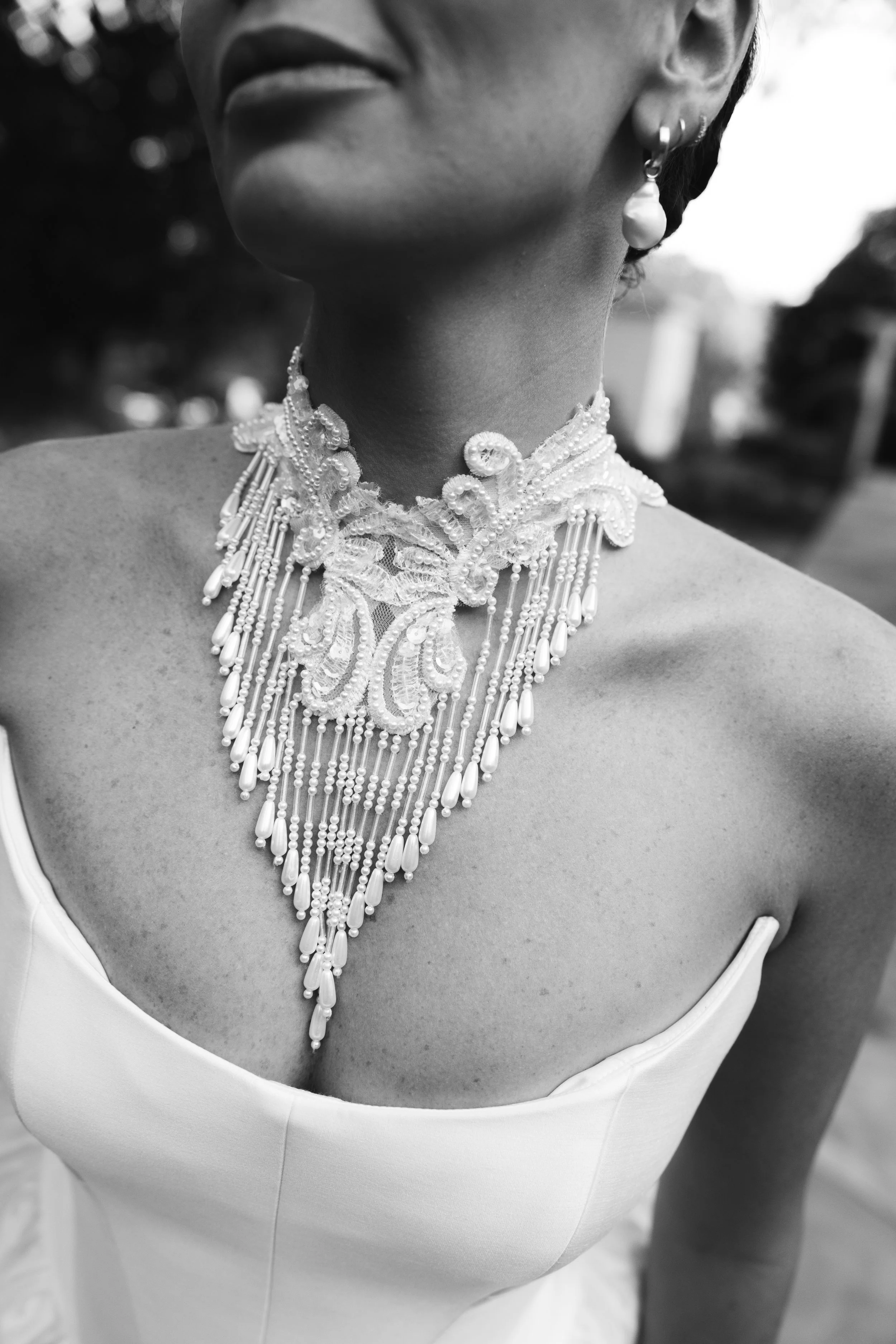 Close-up of a woman wearing an intricate pearl statement necklace, earrings, and a strapless white dress, in black and white photography.