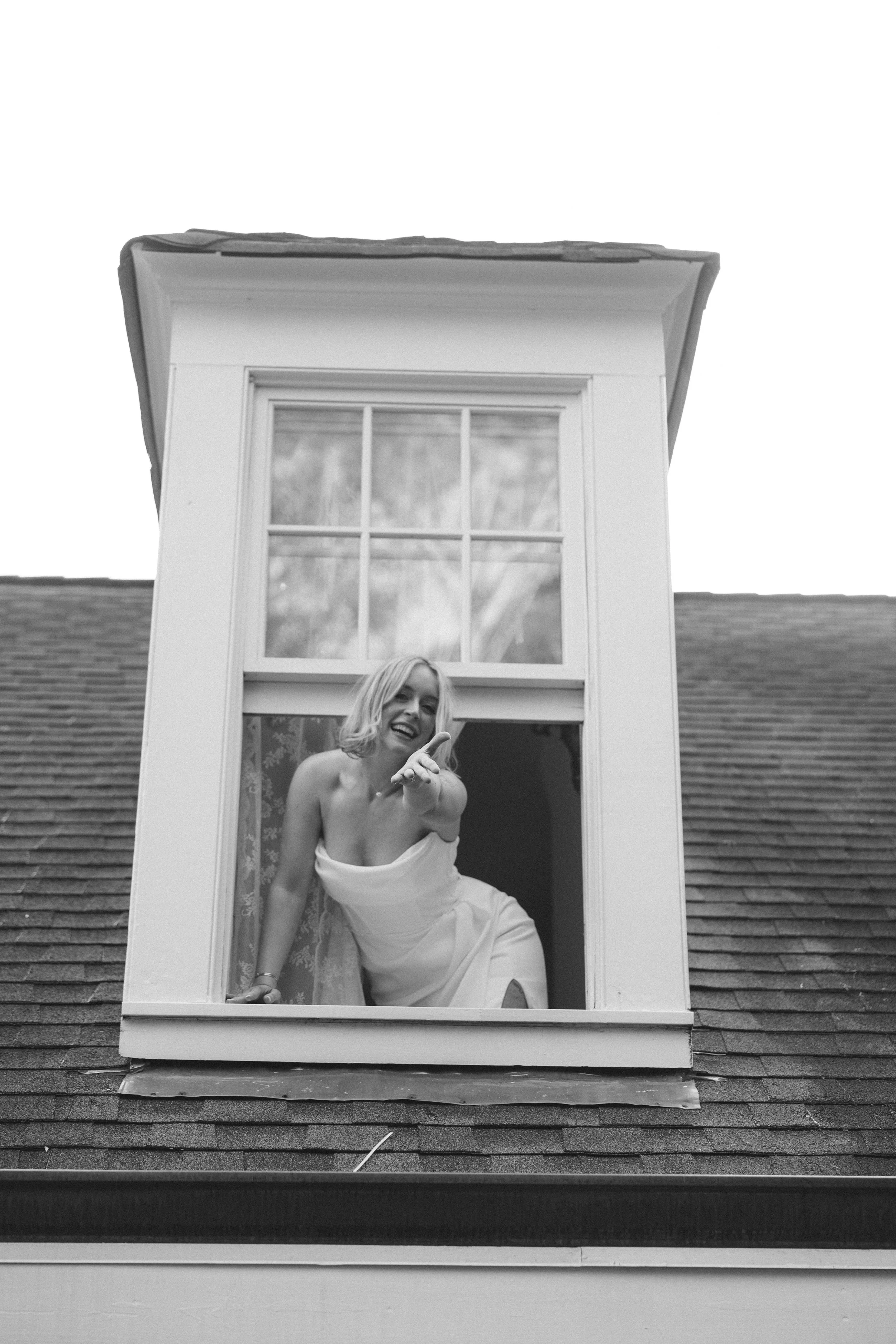 Woman in wedding dress leaning out of a window and pointing at the camera.