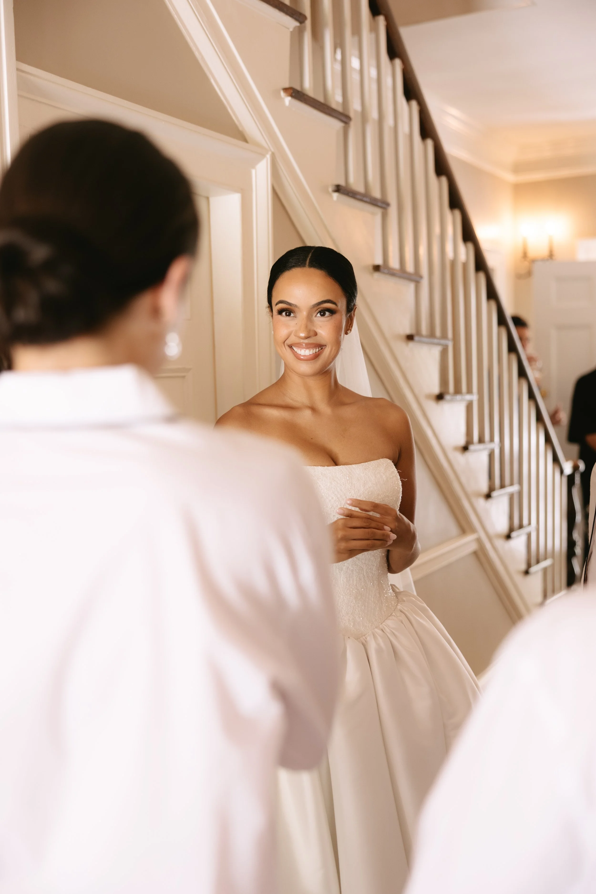 A bride in a strapless wedding gown smiling while talking to another woman, possibly a bridesmaid, in a softly lit, elegant interior with staircase.