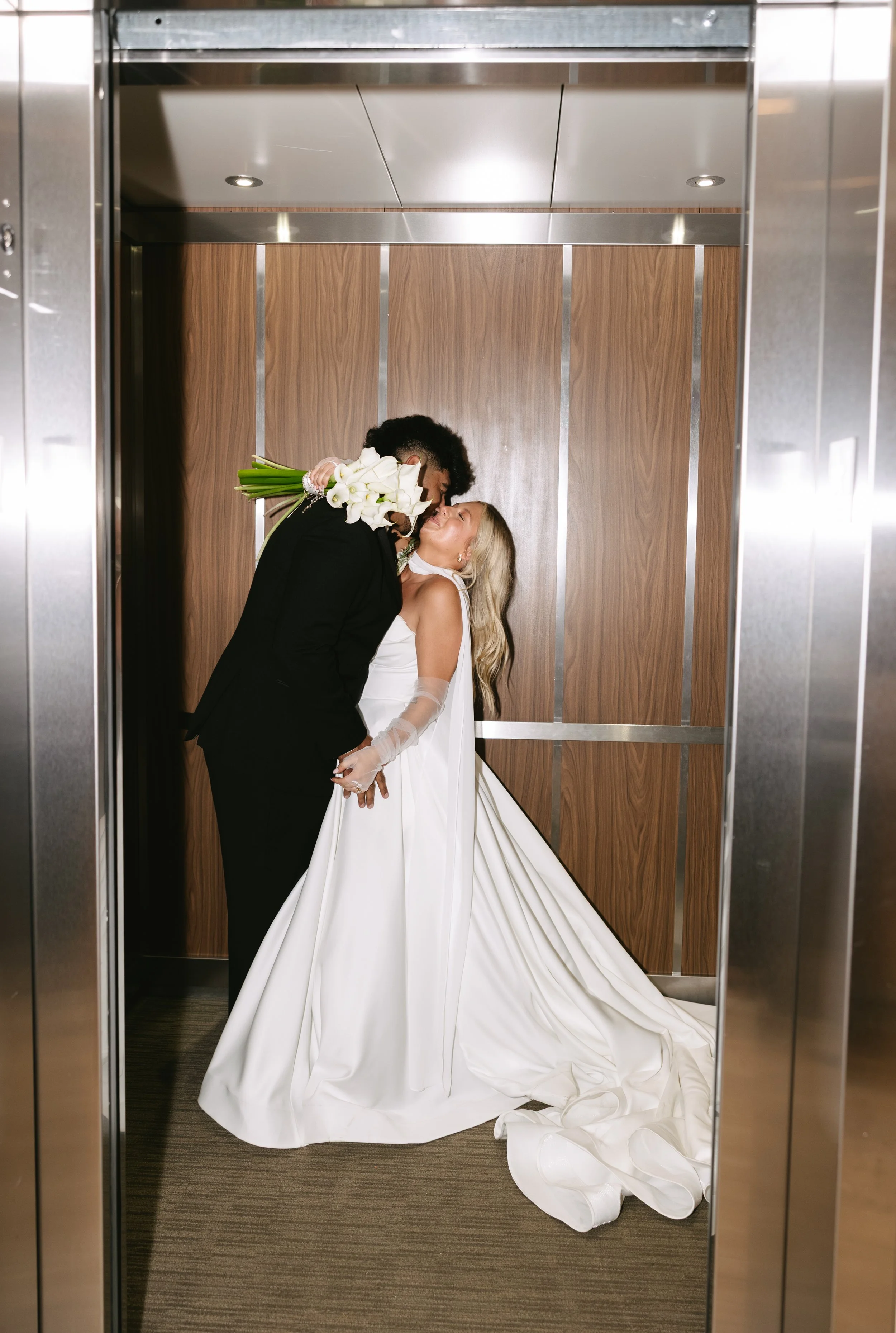 A newlywed couple sharing a kiss in an elevator, with the bride holding a bouquet of white calla lilies.