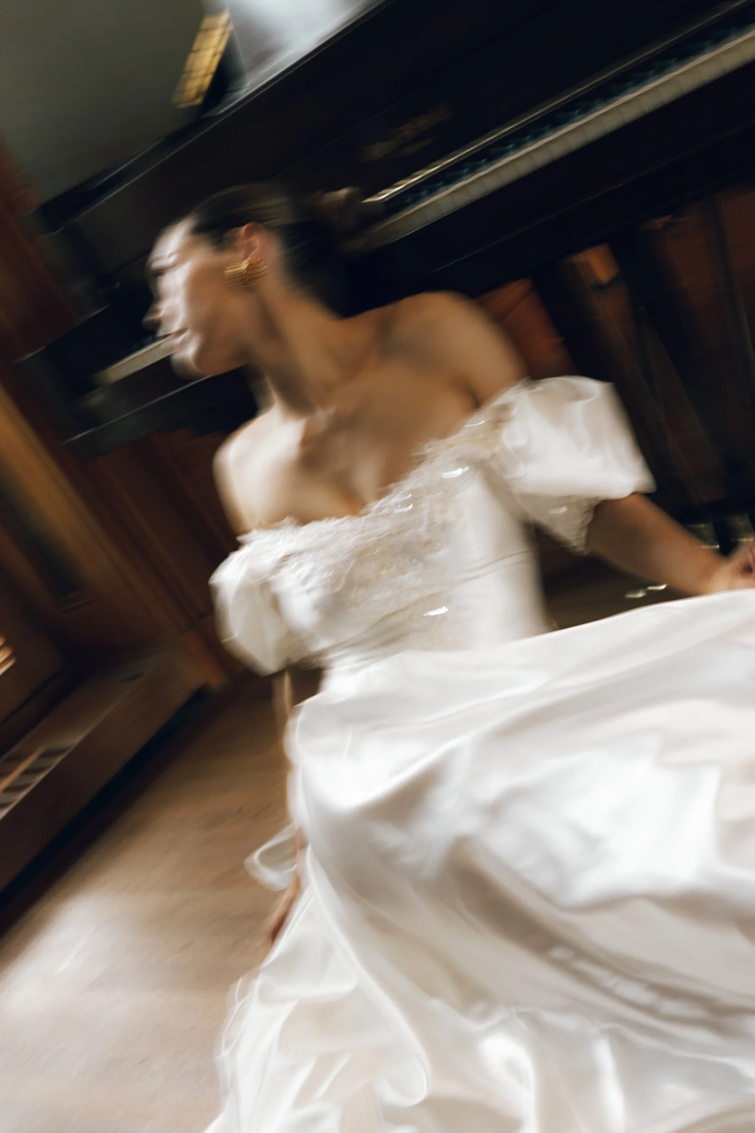 A woman in a white wedding dress is sitting next to a piano, with her head turned to the side, in a room with wooden walls.