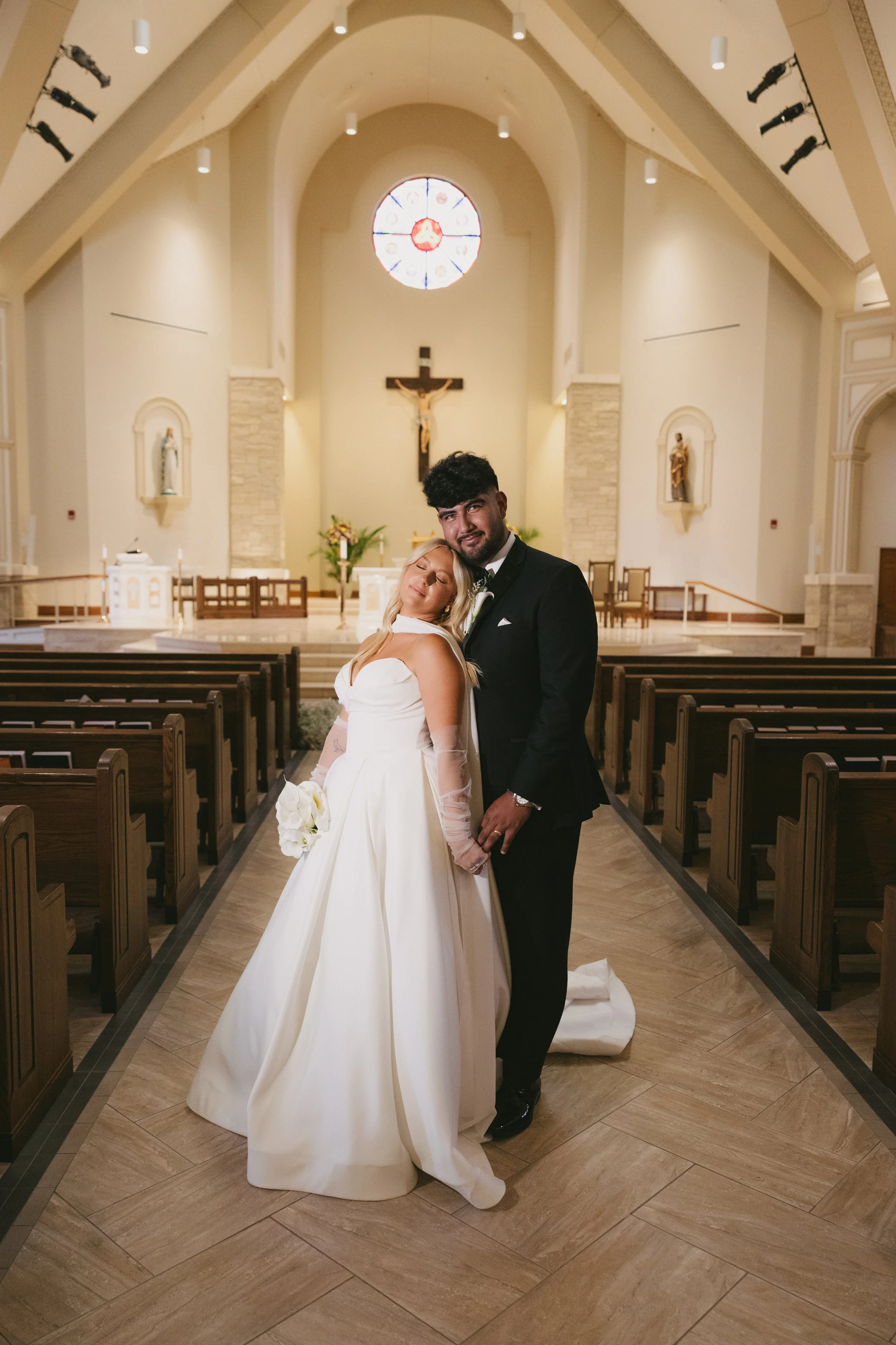Happy bride and groom standing inside a church after their wedding ceremony, with the bride holding a bouquet and the groom in a tuxedo, in front of the altar with a crucifix and religious statues.